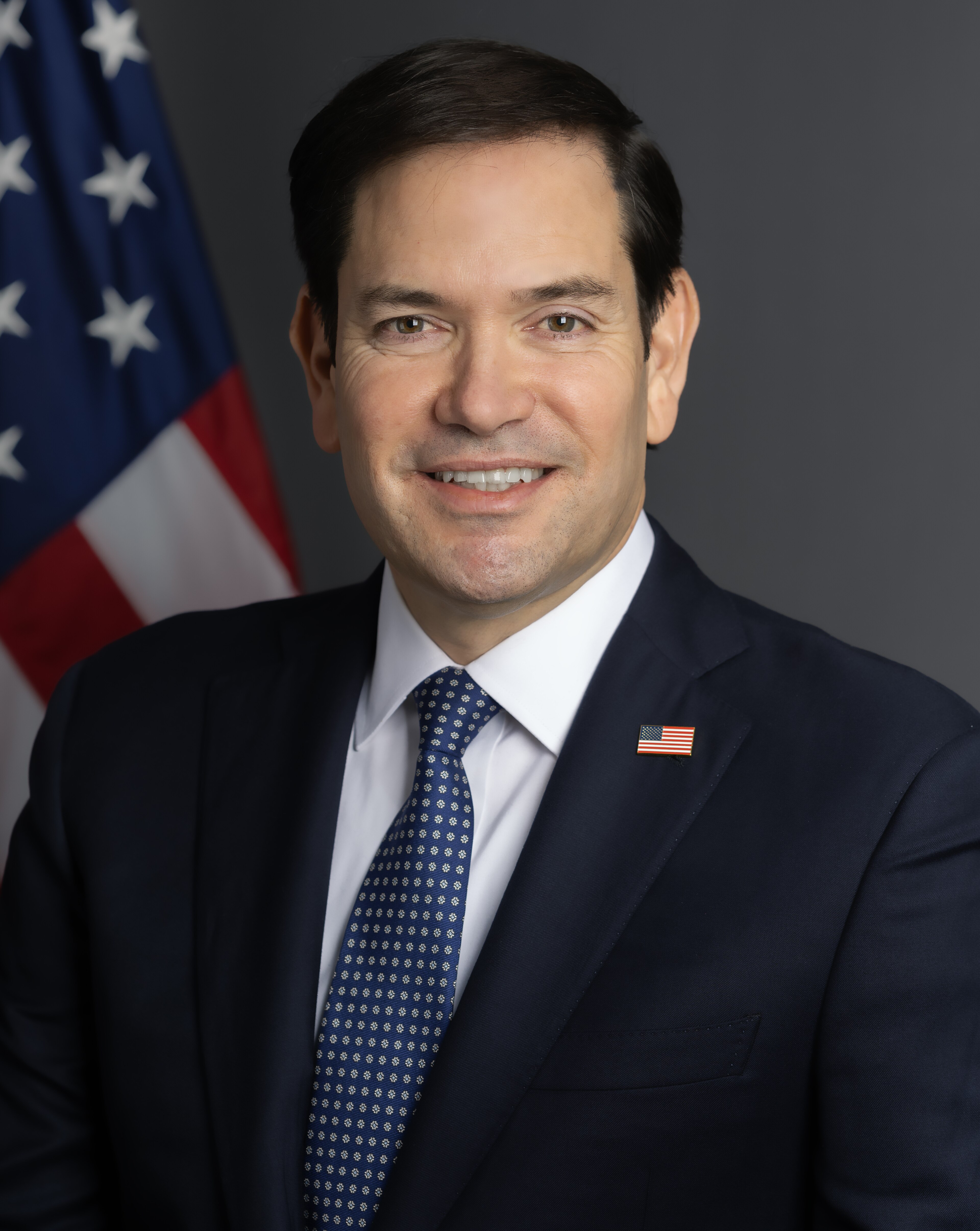 Marco Rubio smiling in his official State Department portrait, US flag lapel pin on a navy suit and blue-and-white patterned tie, 2025