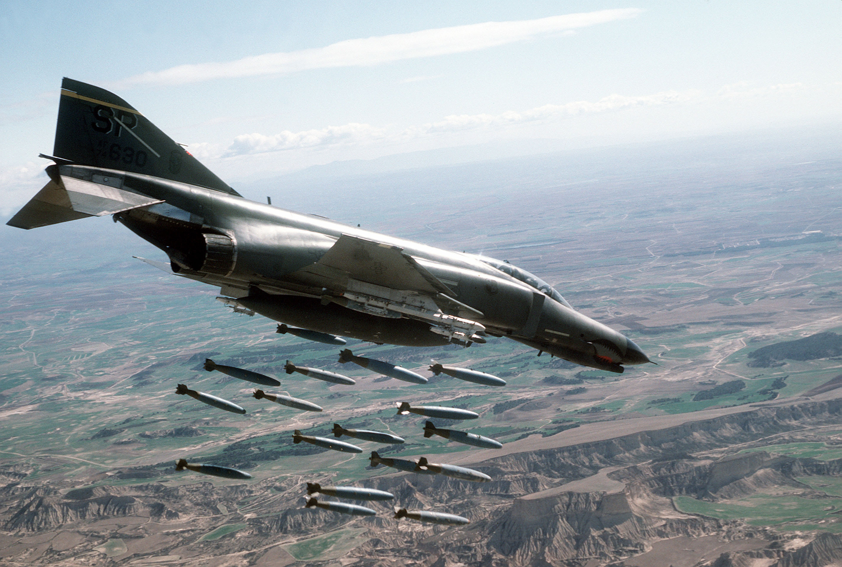 A US Air Force F-4 Phantom releasing a stick of iron bombs above rugged terrain, shark's-mouth nose art and tail number 630, 1985