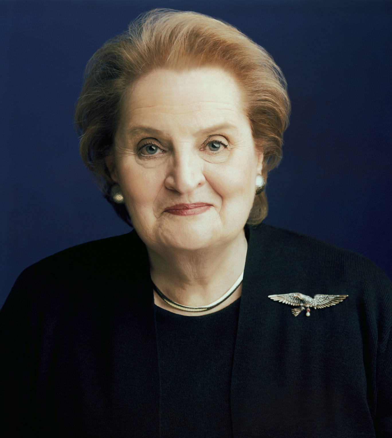 Madeleine Albright in a State Department portrait, black jacket with a silver eagle brooch on the lapel against a deep blue backdrop, 2000