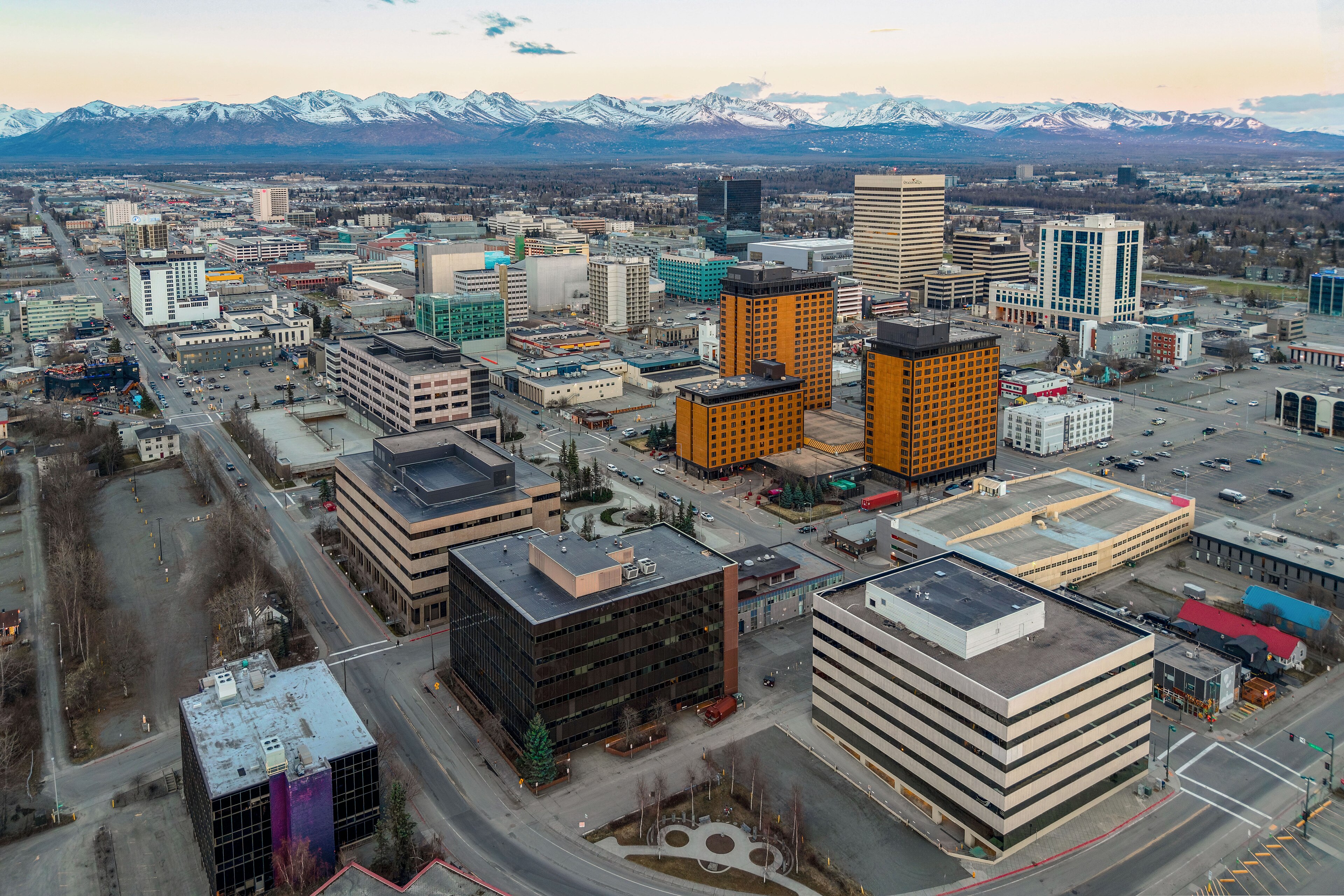 Aerial view of downtown Anchorage at dusk, low office blocks and orange hotel towers against the snow-capped Chugach mountains, 2015