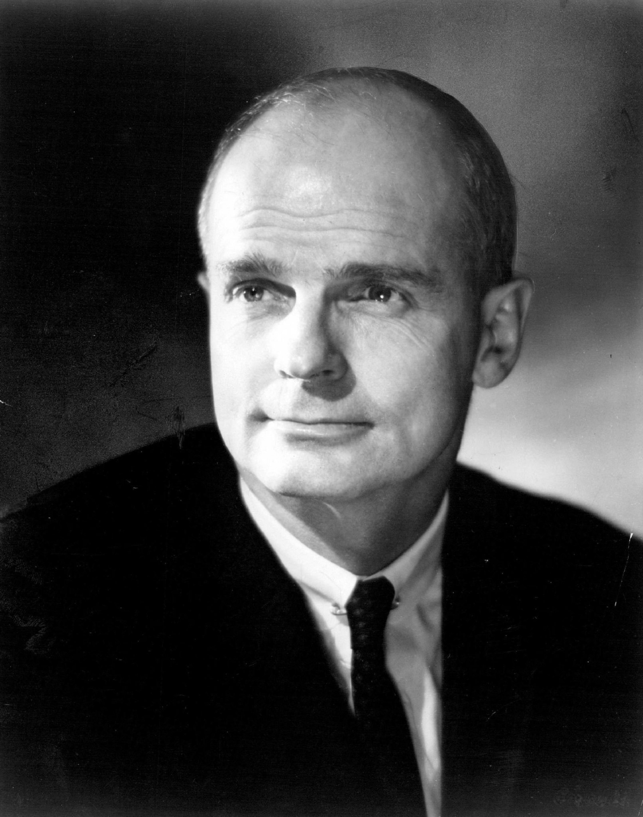 William Proxmire in a black-and-white official portrait, bald head above a dark suit and thin knit tie against a soft grey wash, 1978