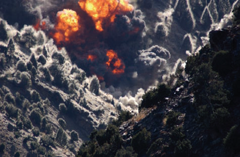 An orange fireball detonating on the wooded slope of a Tora Bora ridge, smoke rolling out through the trees, 2001