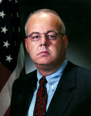 Cofer Black in an official portrait, bald head and wire-frame glasses over a red-and-blue striped tie under a dark suit against the US flag, 2001