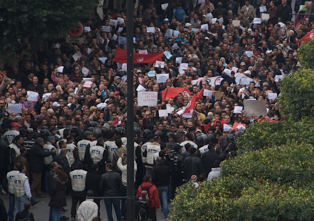 A crowd of Tunisians waving red-and-white Tunisian flags at a downtown Tunis protest, police in white bibs at the front line, 2011