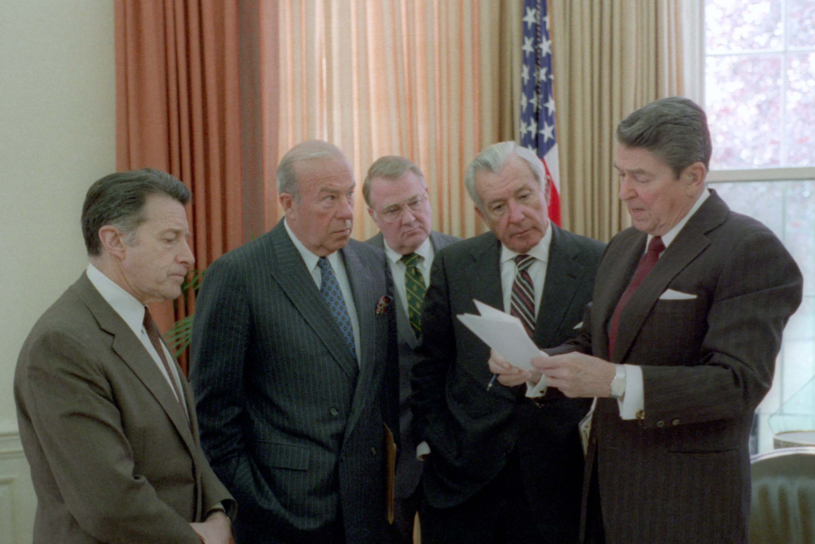 Ronald Reagan reading from a document in the Oval Office to Caspar Weinberger, George Shultz, Ed Meese and William Casey standing before the drapes, 1985