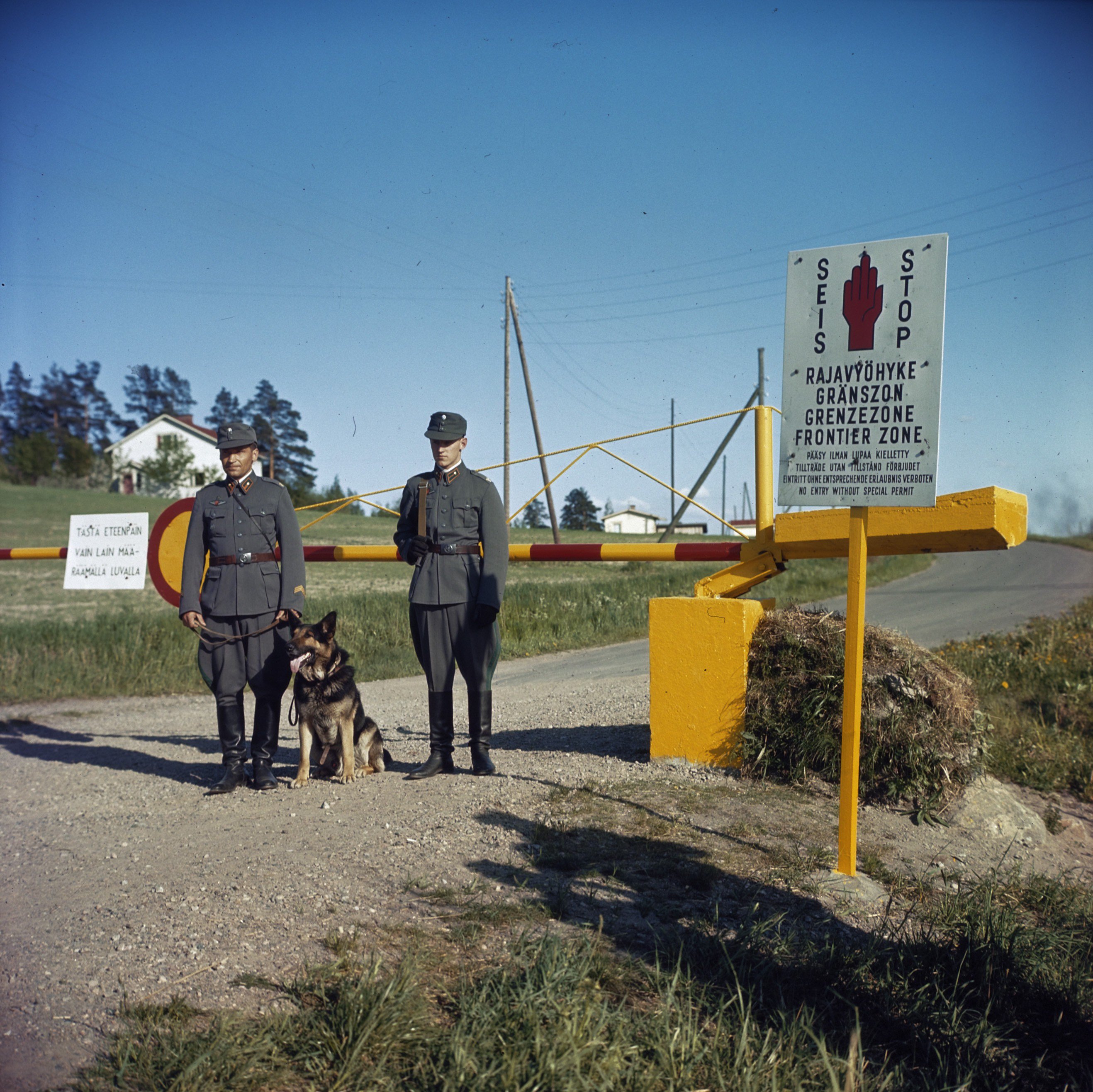 Two Finnish border guards with a shepherd dog at a yellow-and-red barrier under a Frontier Zone sign, 1960