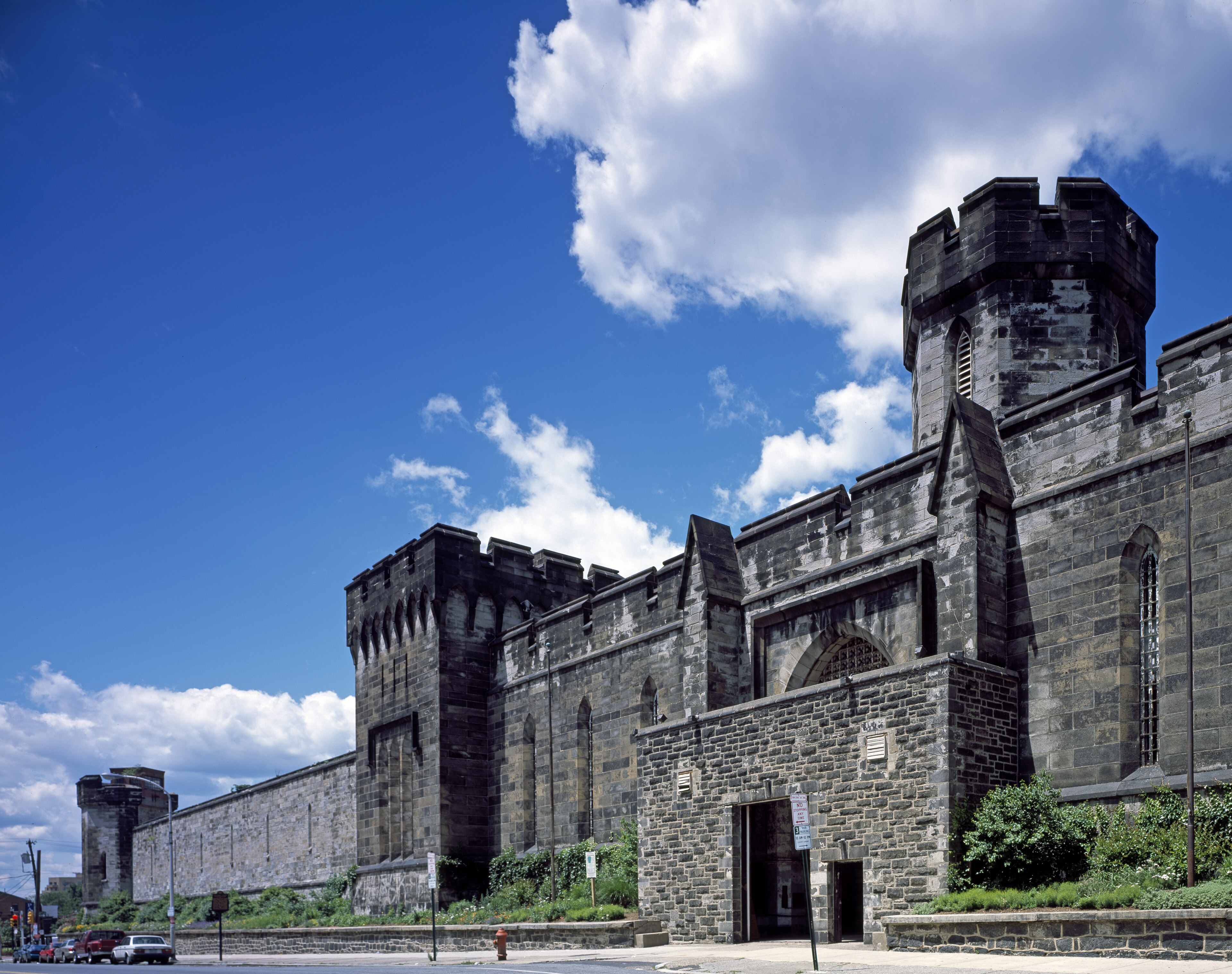 Eastern State Penitentiary in Philadelphia, neo-Gothic stone turrets rising over the perimeter wall under a blue summer sky, 1829