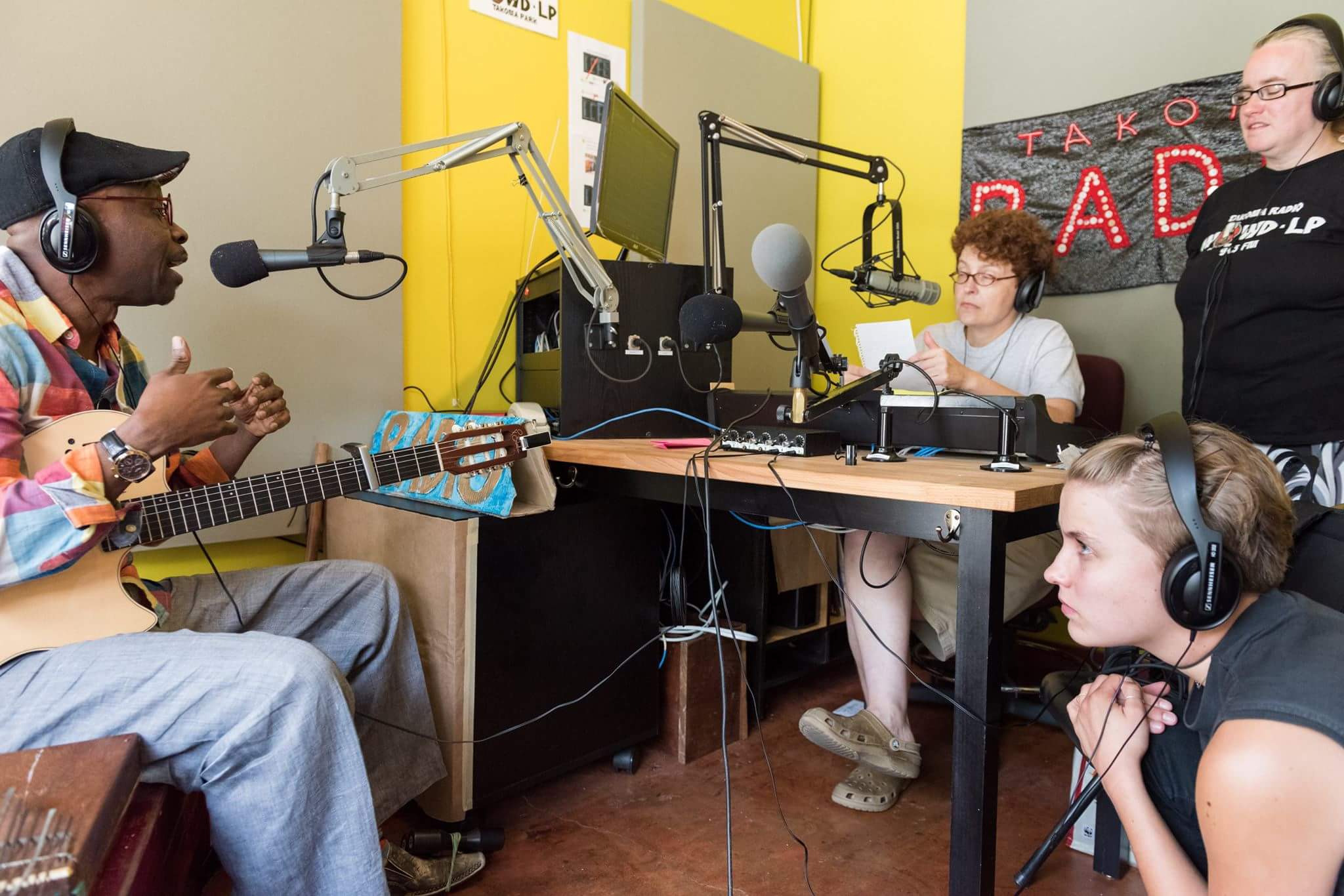A community radio interview in progress at WOWD-LP Takoma Park, a guest musician on acoustic guitar at the microphone with two producers seated at the console, 2015