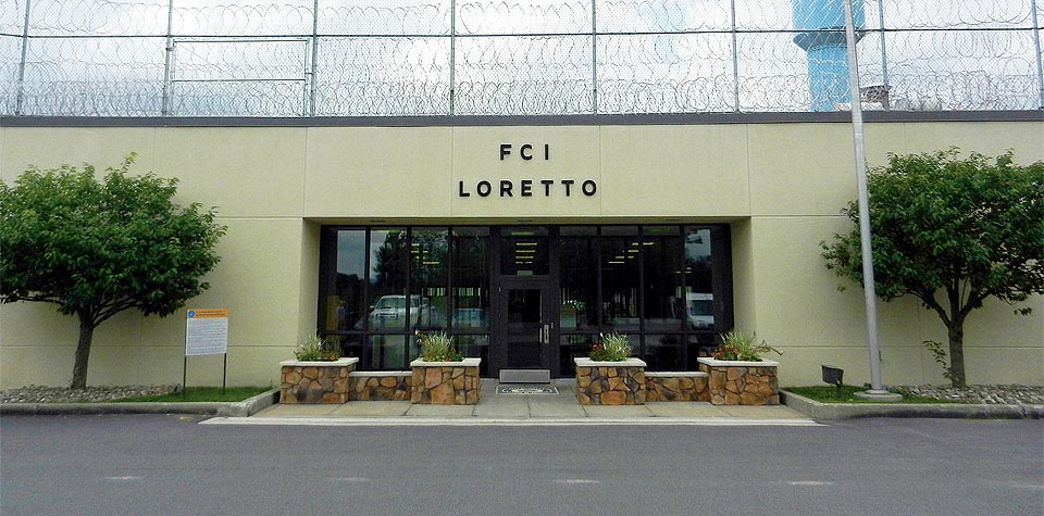The gated entrance to FCI Loretto with a flowering magnolia in the foreground and a US flag on the pole above, 2013