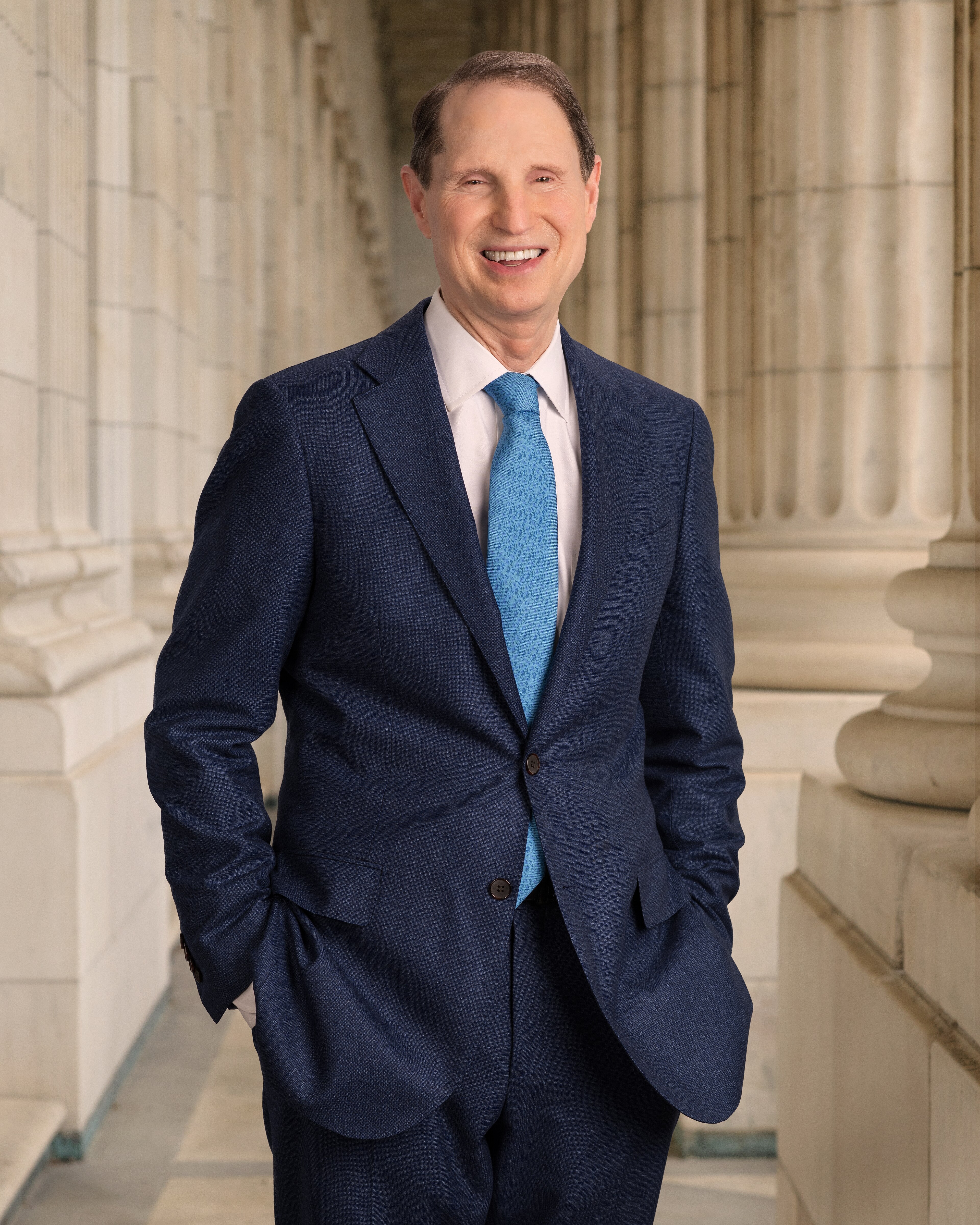 Ron Wyden smiling in a candid portrait, hands in the pockets of a navy suit and light-blue paisley tie against a limestone Capitol colonnade, 2015