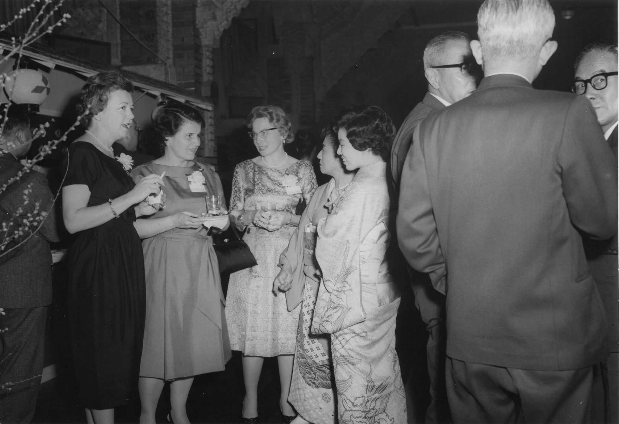 A black-and-white cocktail-party photograph, American women in evening dresses and Japanese women in kimono speaking beside a group of grey-suited men, 1962
