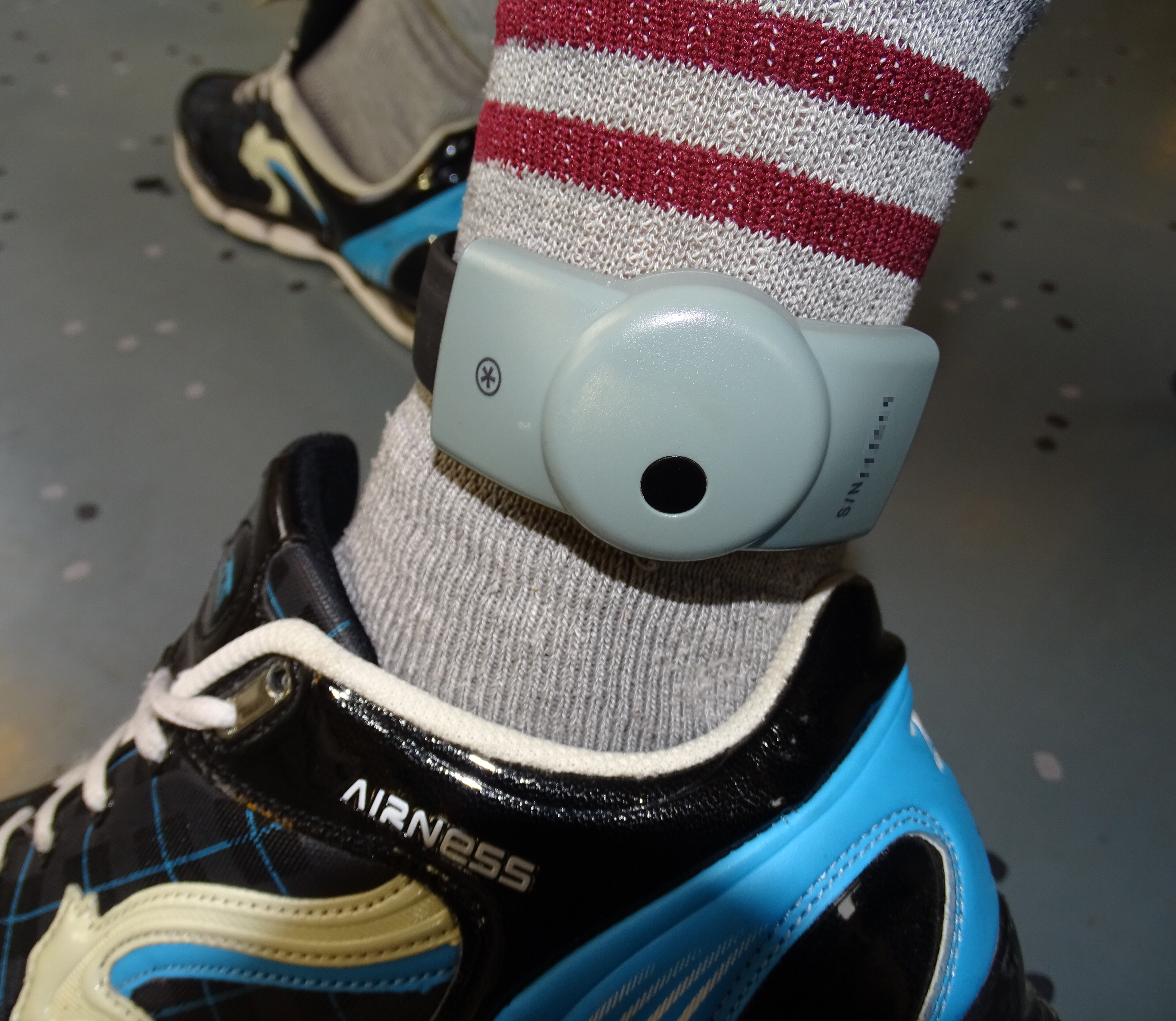 A grey electronic ankle monitor fastened over a striped sock above a black-and-blue running shoe, 2015