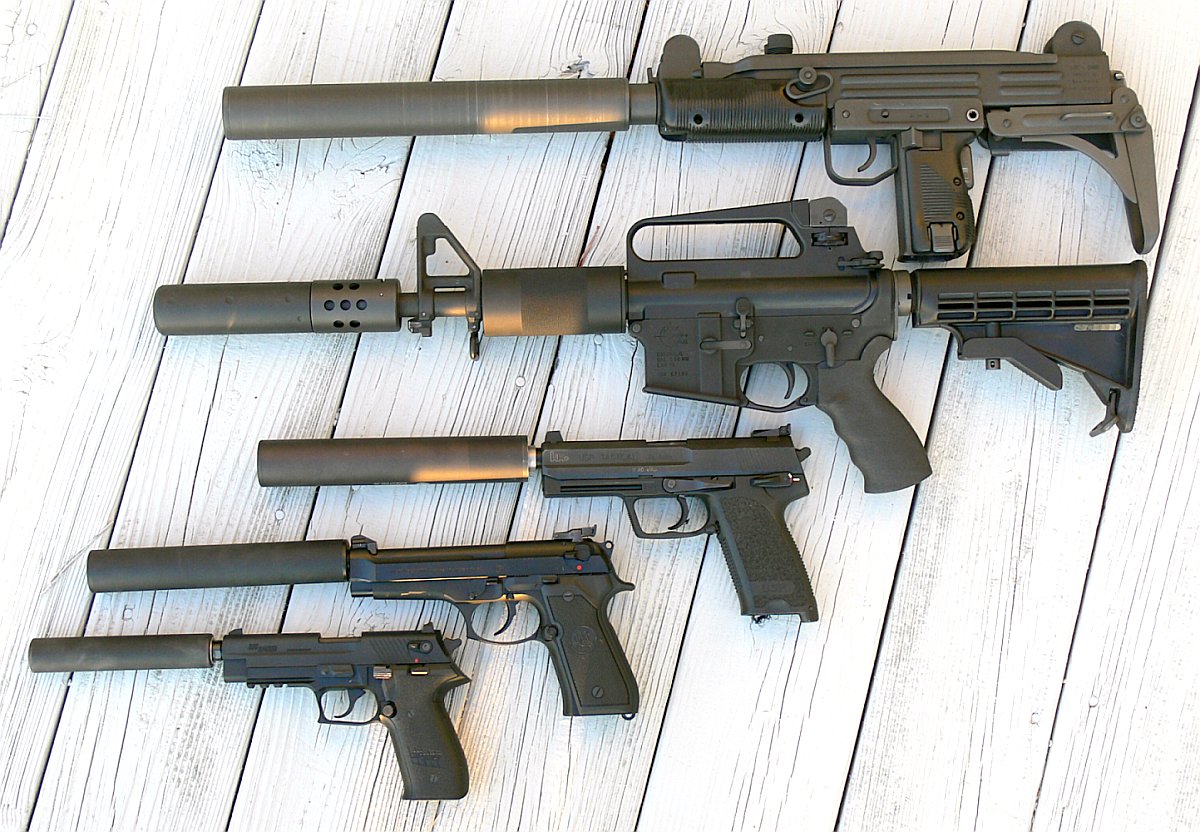 A row of suppressed firearms, from an Uzi and M4 carbine down to three pistols with silencers, laid on a wooden deck, 2010