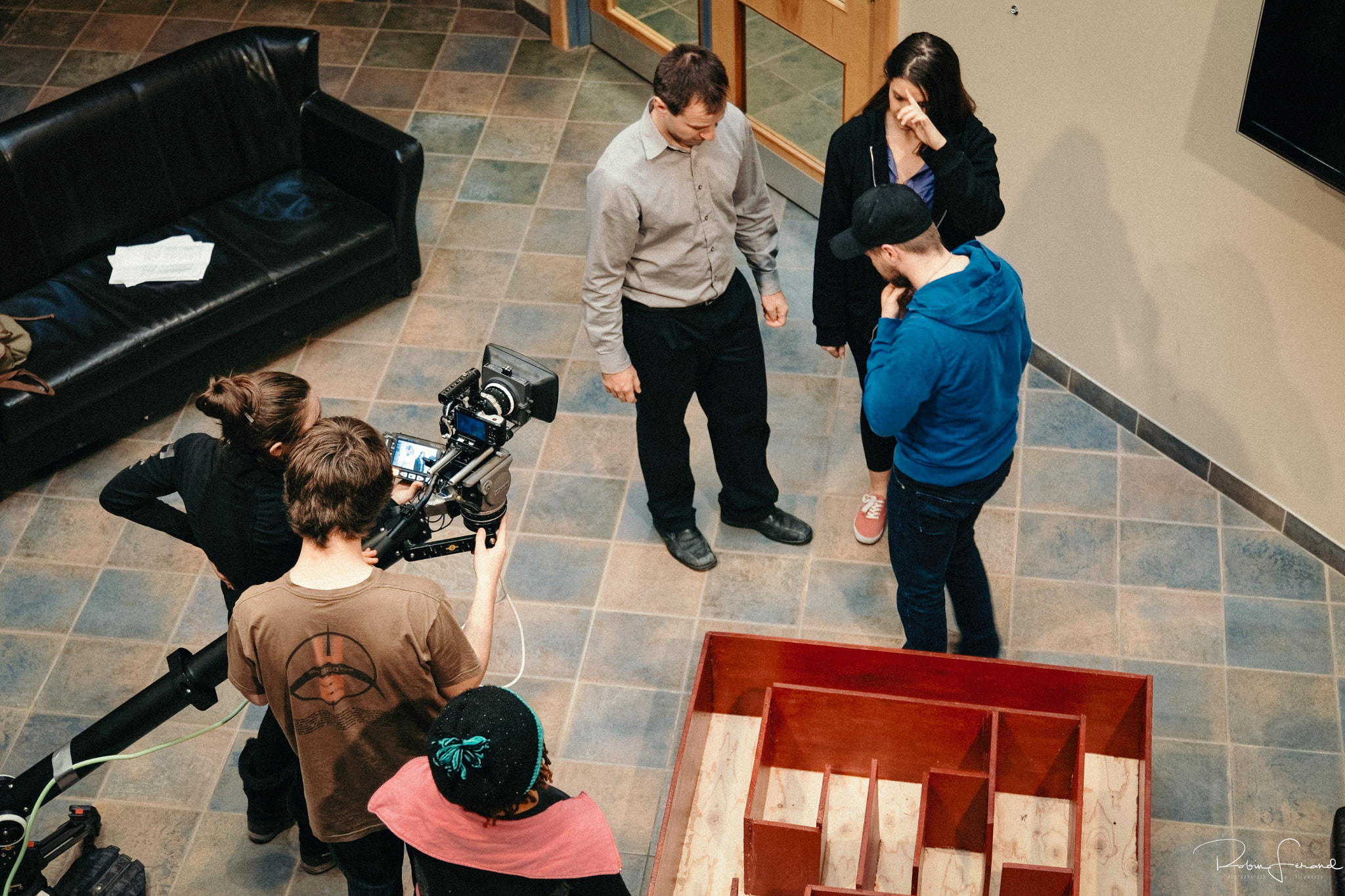 An overhead view of a small film crew rehearsing a scene between two actors beside a leather couch, camera operator crouched with a rig, 2020