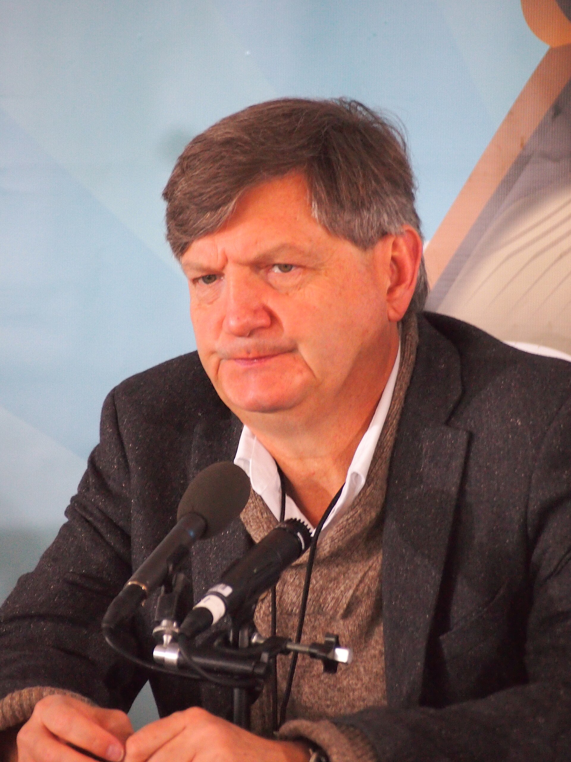 James Risen speaking into a pair of microphones in a grey jacket and open collar, 2015