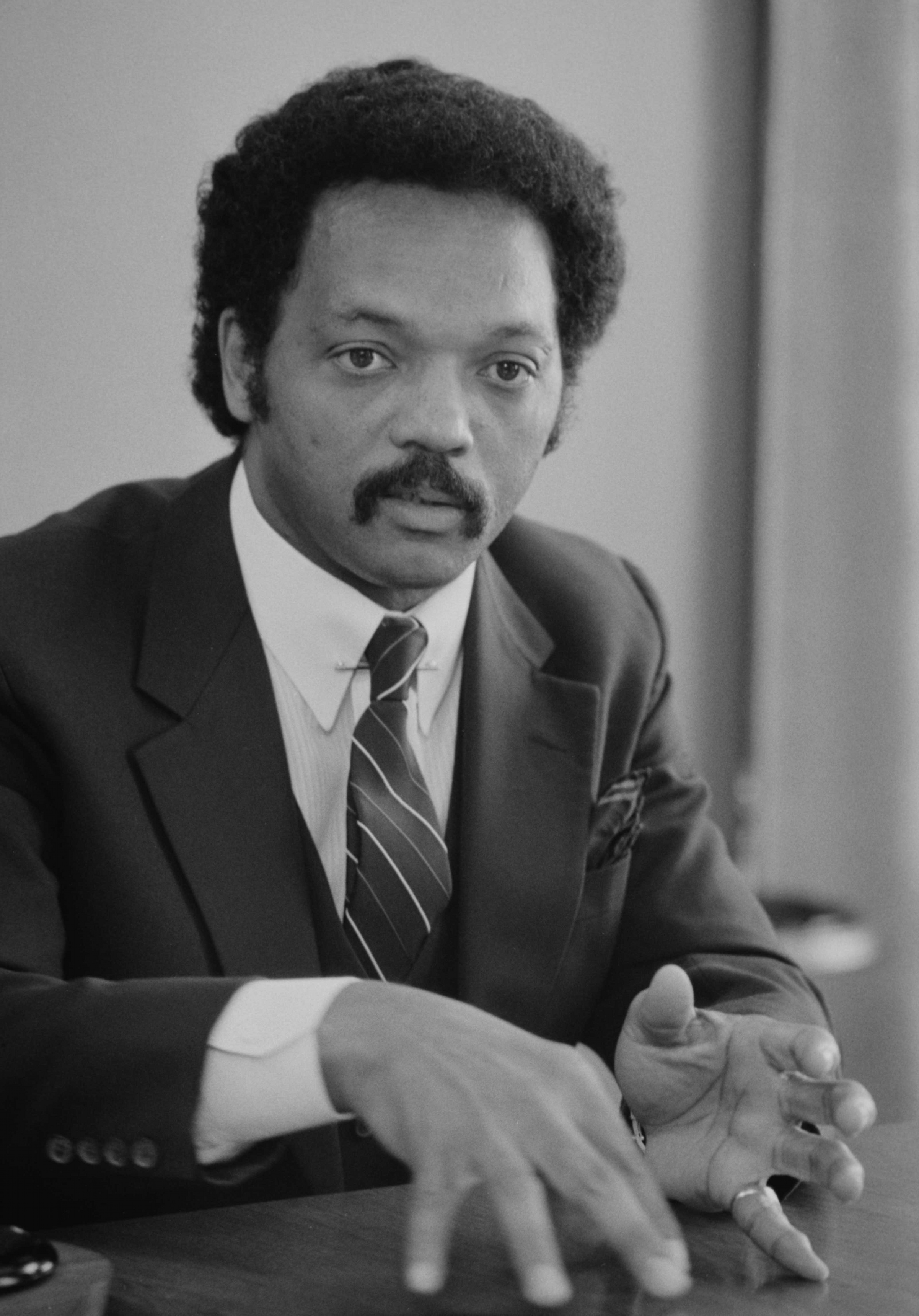 Jesse Jackson in a black-and-white portrait, hands folded on a wooden table under a dark blazer and striped tie, 1980