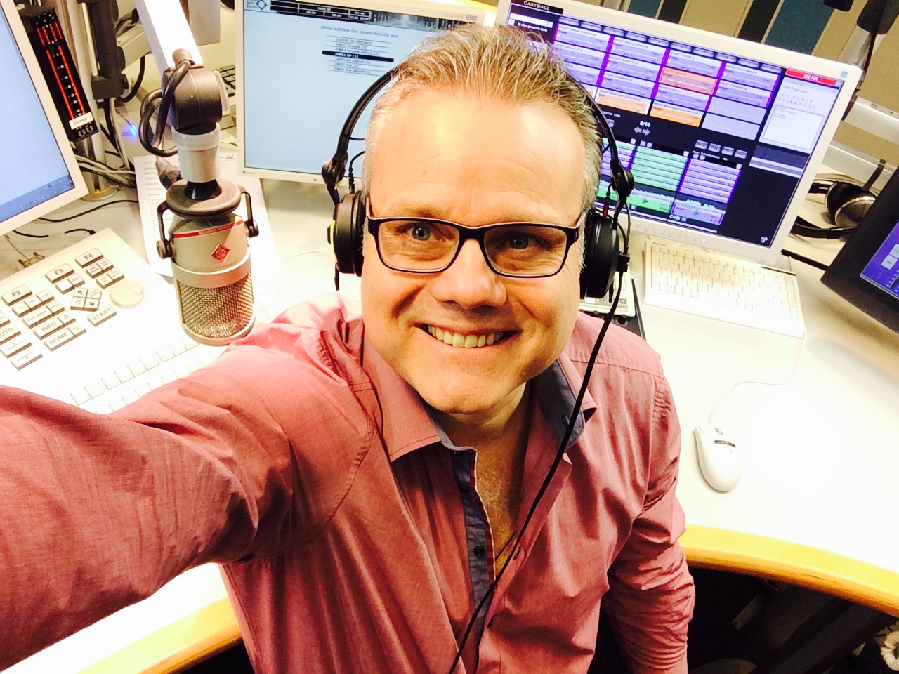 A radio host in a coral shirt taking a selfie at his studio microphone under a set of headphones, station software glowing behind him, 2015