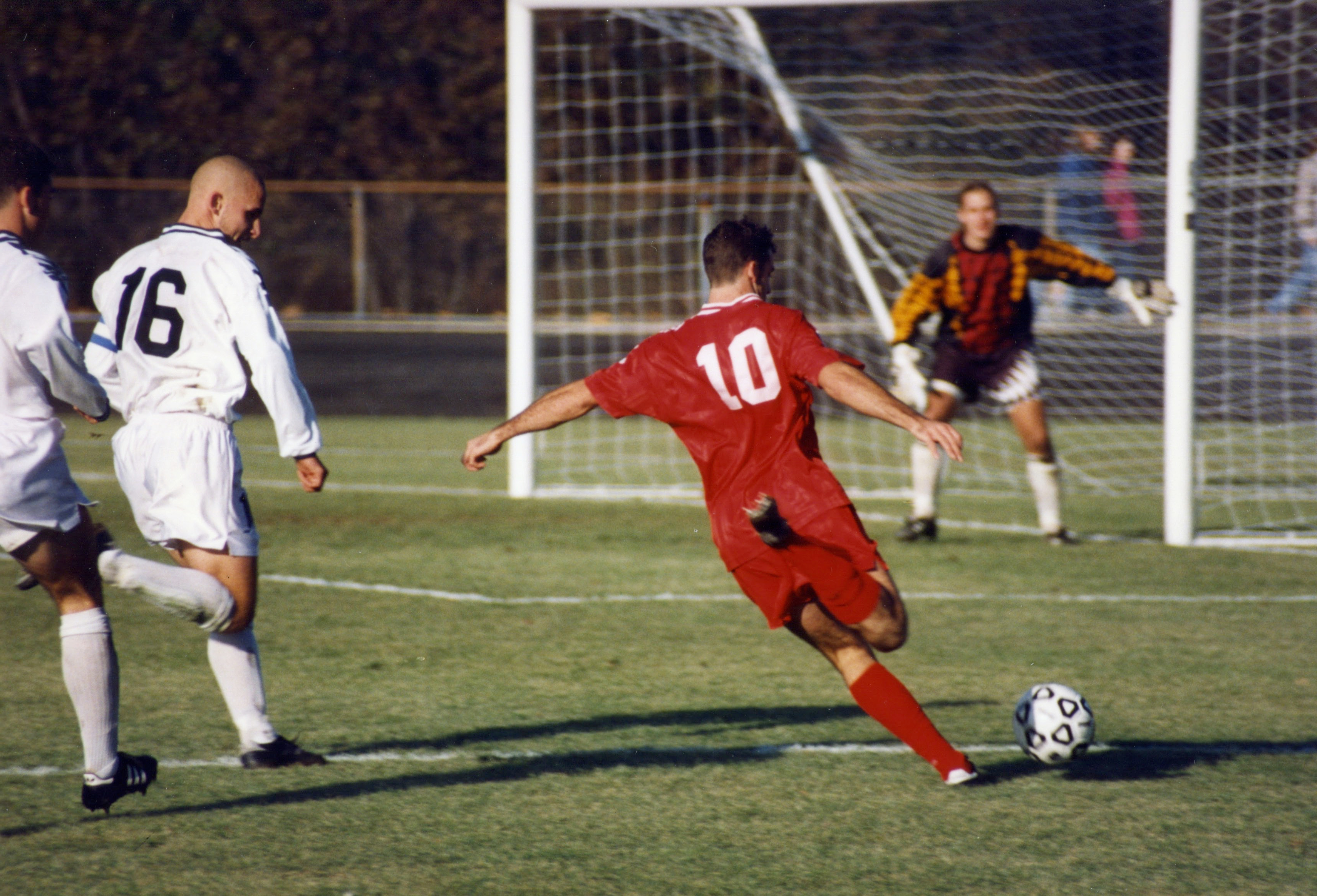 A red-shirted number 10 striker striding into a shot on goal as a white defender closes and the keeper spreads himself across the net, 2015