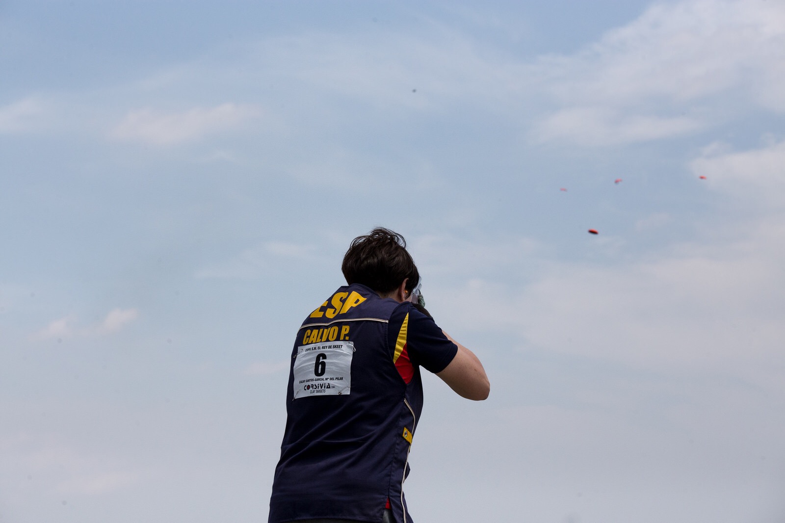 A skeet shooter in an ESP club vest lifting a shotgun to break a pair of clay targets against a pale sky, 2015
