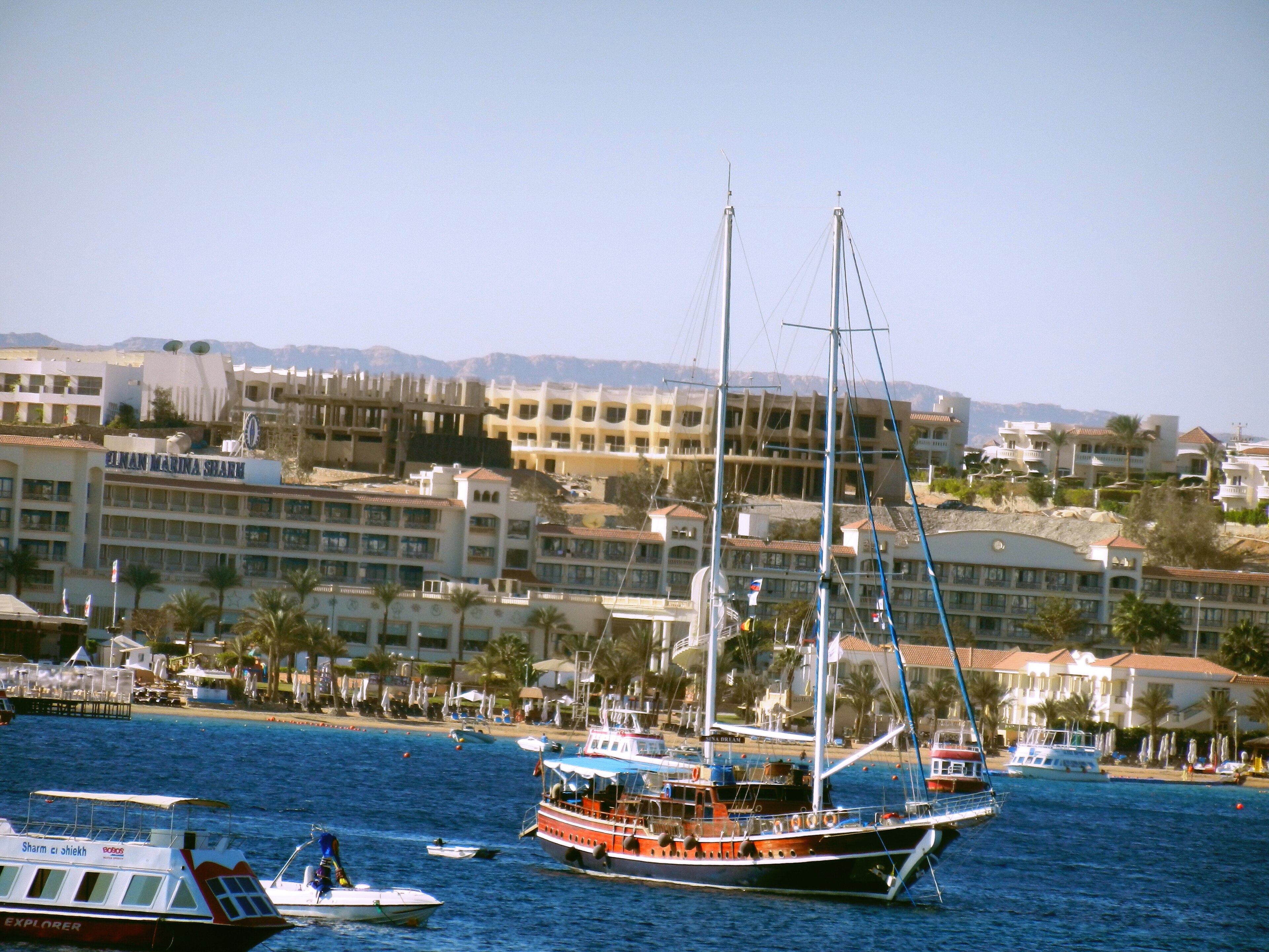 A two-masted red-and-blue tourist schooner and a Sharm el-Sheikh dive boat moored off the Sinan Marina Sharm resort under desert hills, 2010