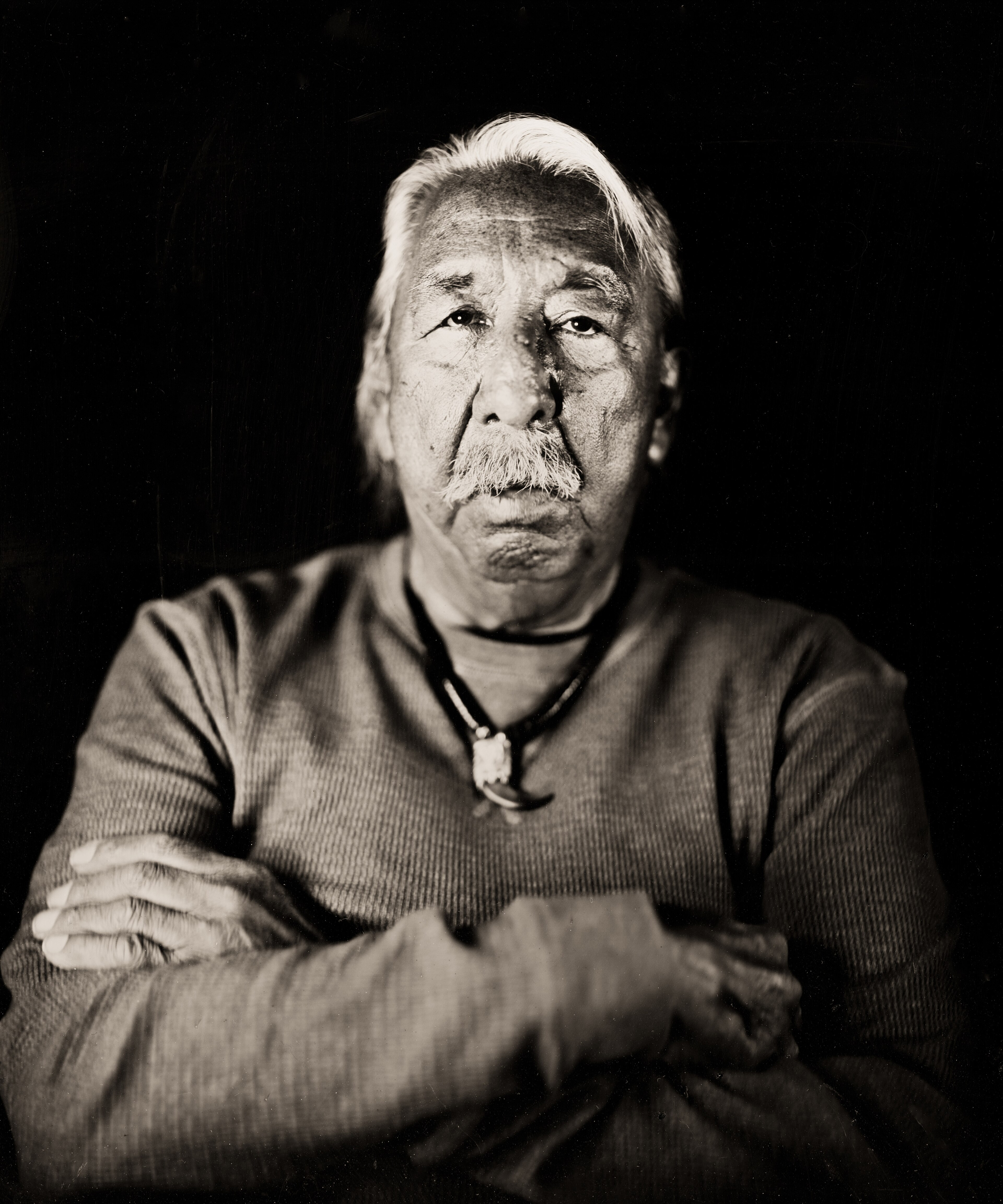 Leonard Peltier in a sepia arms-crossed portrait, white hair swept back over a ribbed grey shirt and a bear-claw pendant, 2015