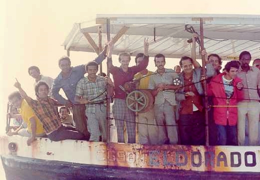 Cuban Mariel migrants crammed on the deck of the shrimper Eldorado, one hoisting a sign at the bow as the boat rides low in the Caribbean, 1980