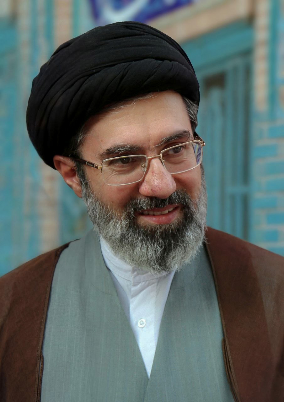 Mojtaba Khamenei smiling in a black turban and grey beard behind wire-frame glasses, a green vest over a brown robe against a turquoise-tiled wall, 2018