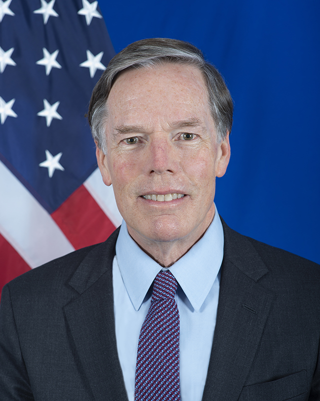 Nicholas Burns smiling in an official State Department portrait, US flag behind him and a red-and-blue chequered tie under a dark suit, 2021
