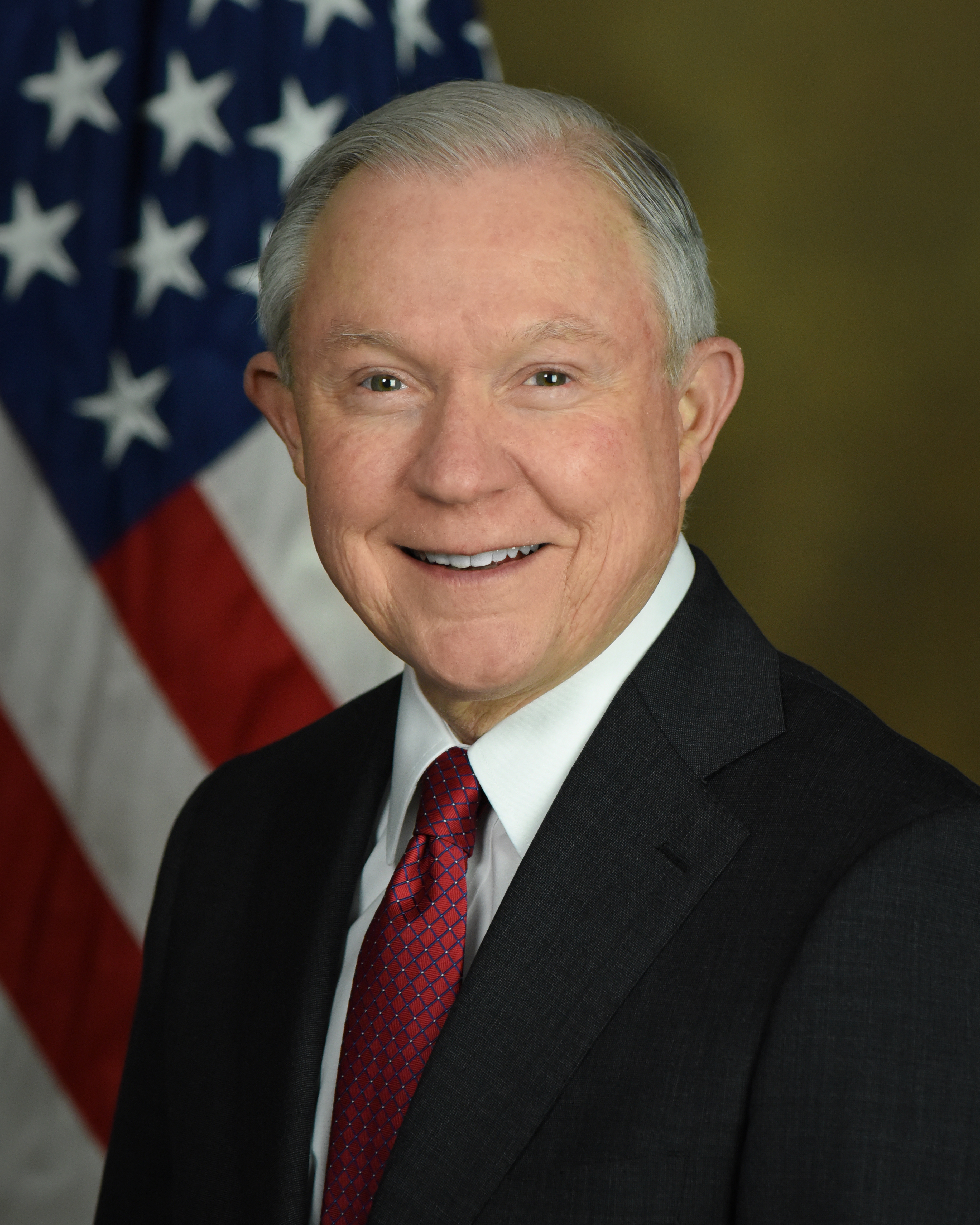 Jeff Sessions smiling in his official Justice Department portrait, white hair over a red diamond-patterned tie under a dark suit against the US flag, 2017