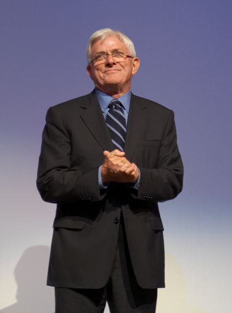 Phil Donahue clasping his hands on stage in a dark suit and striped tie, 2010