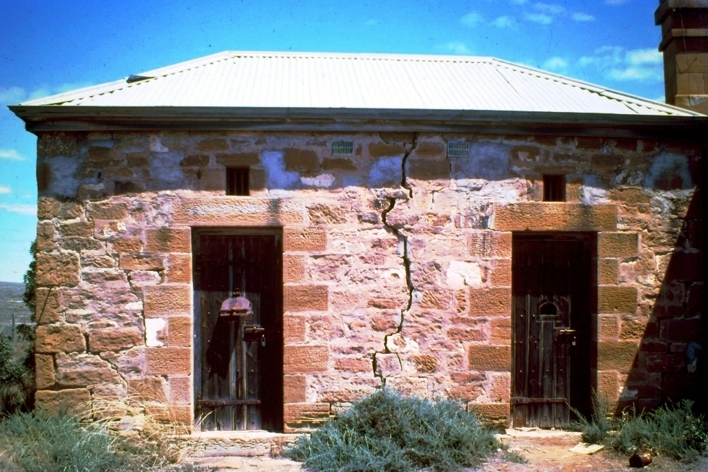An old sandstone jailhouse with two wooden doors under a corrugated metal roof, a long crack running down the facade, 1900
