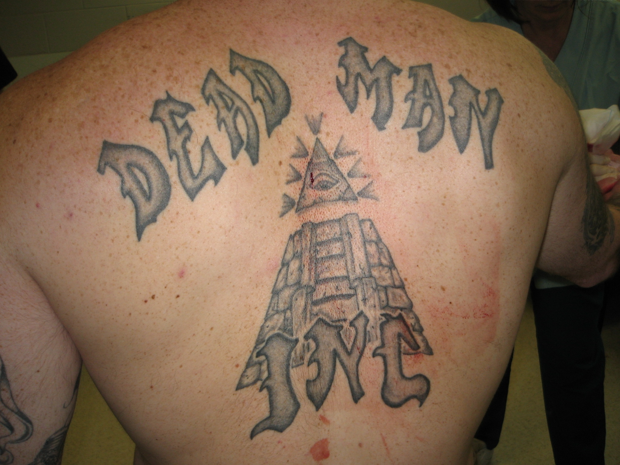 A "DEAD MAN INC" gothic tattoo curved above an all-seeing eye and pyramid across a freckled prisoner's upper back, 2010