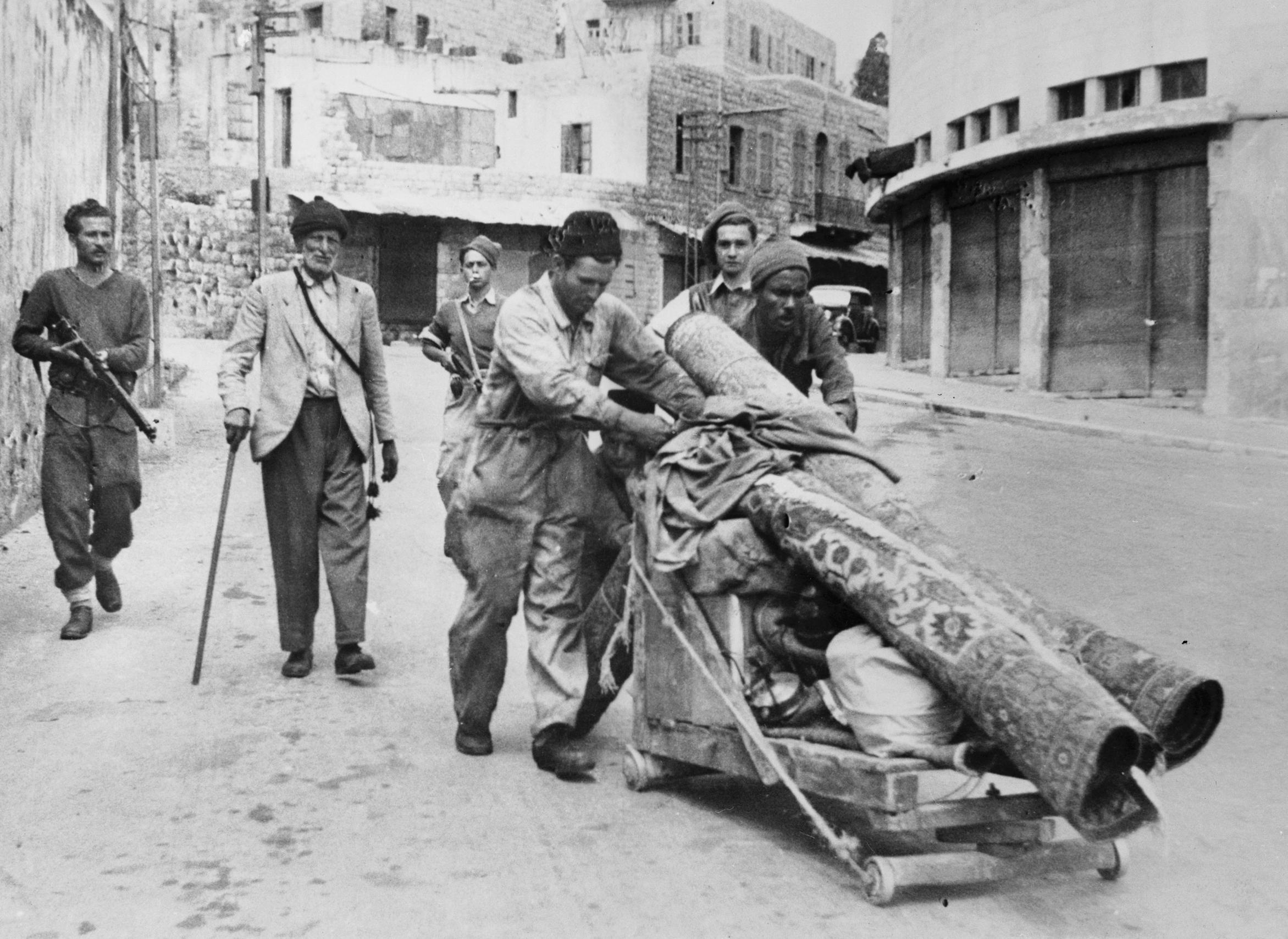 Palestinians pushing a handcart of rolled carpets and belongings down a Haifa street under armed Haganah escort during the Nakba, 1948