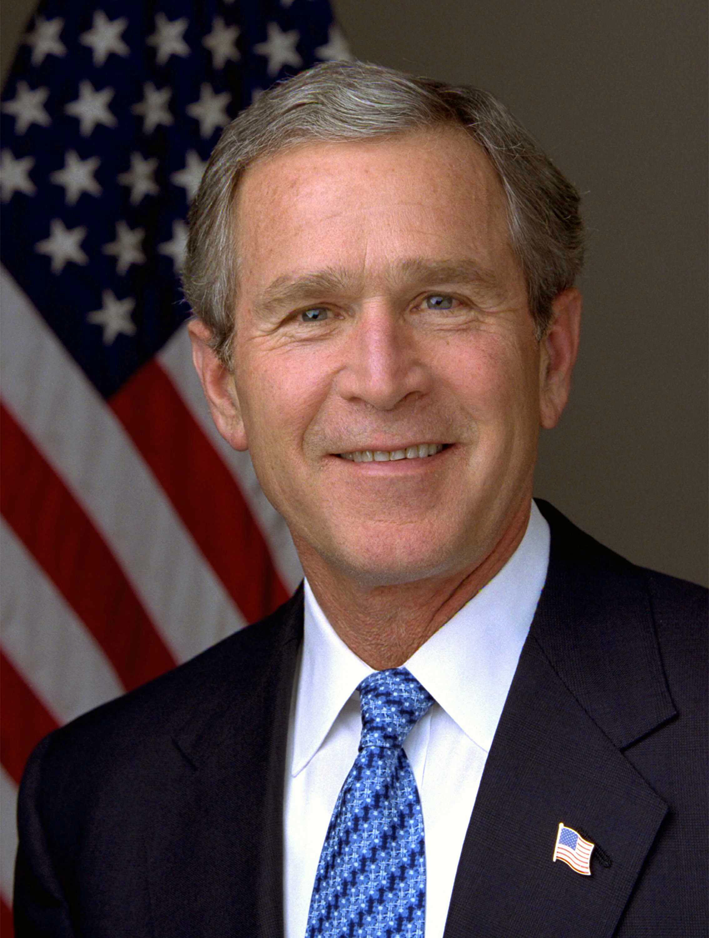 George W. Bush smiling in his official presidential portrait, US flag lapel pin over a blue patterned tie under a dark suit against the US flag, 2001