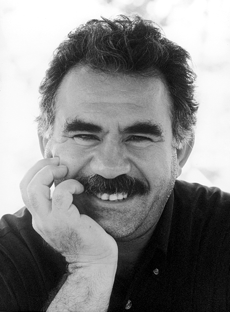 Abdullah Ocalan smiling in a black-and-white outdoor portrait, thick mustache above a hand resting at his chin, 1999