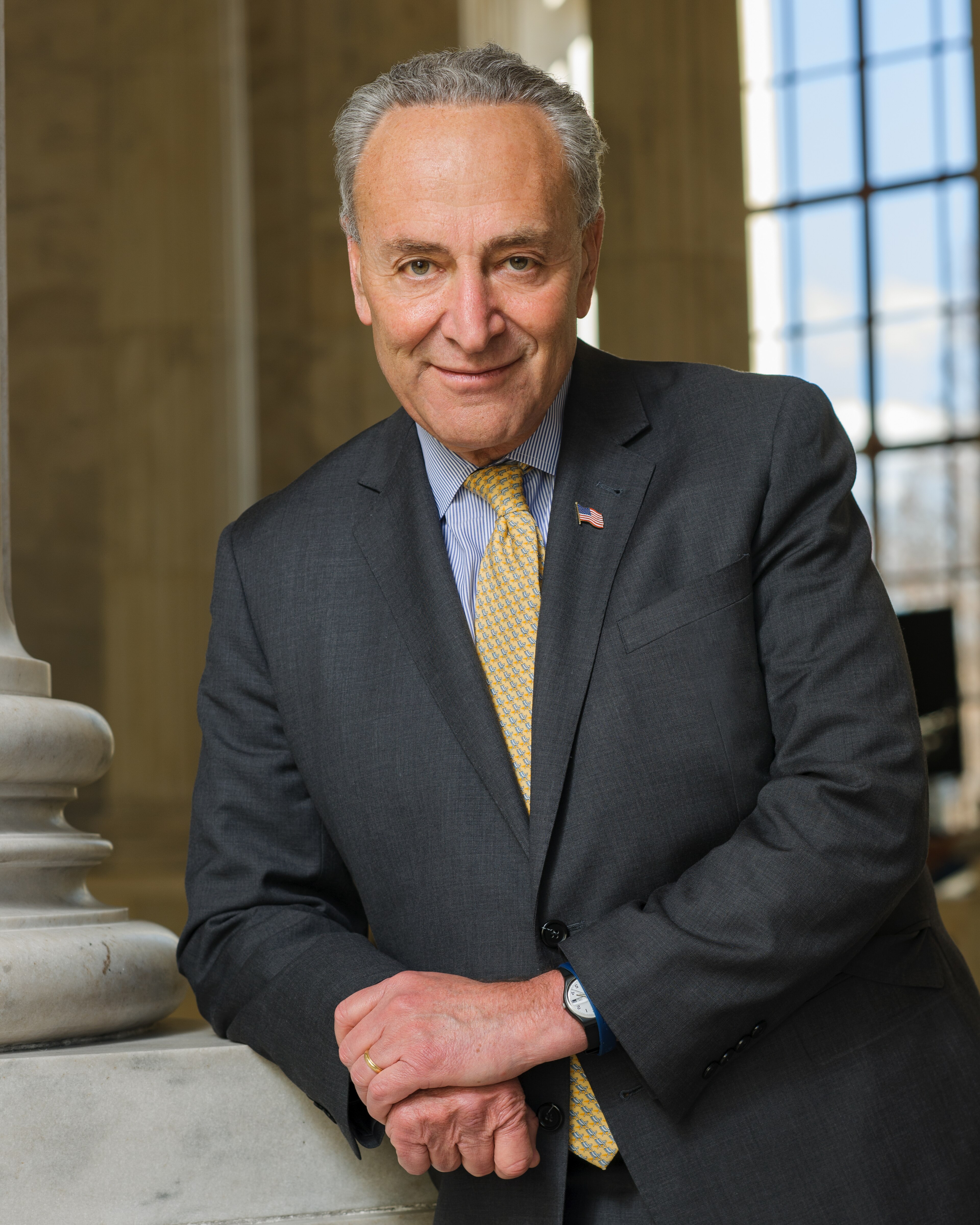 Chuck Schumer leaning on a marble balustrade inside the Capitol, grey suit and yellow paisley tie under a US flag lapel pin, 2015