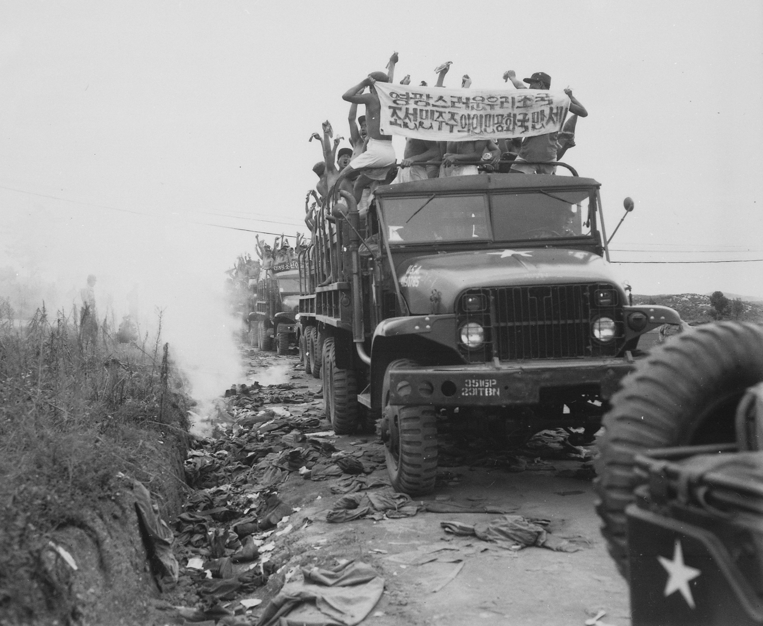 A black-and-white photograph of released Korean War prisoners riding stripped to the waist on top of US Army trucks, a Korean-script banner raised above them, 1953