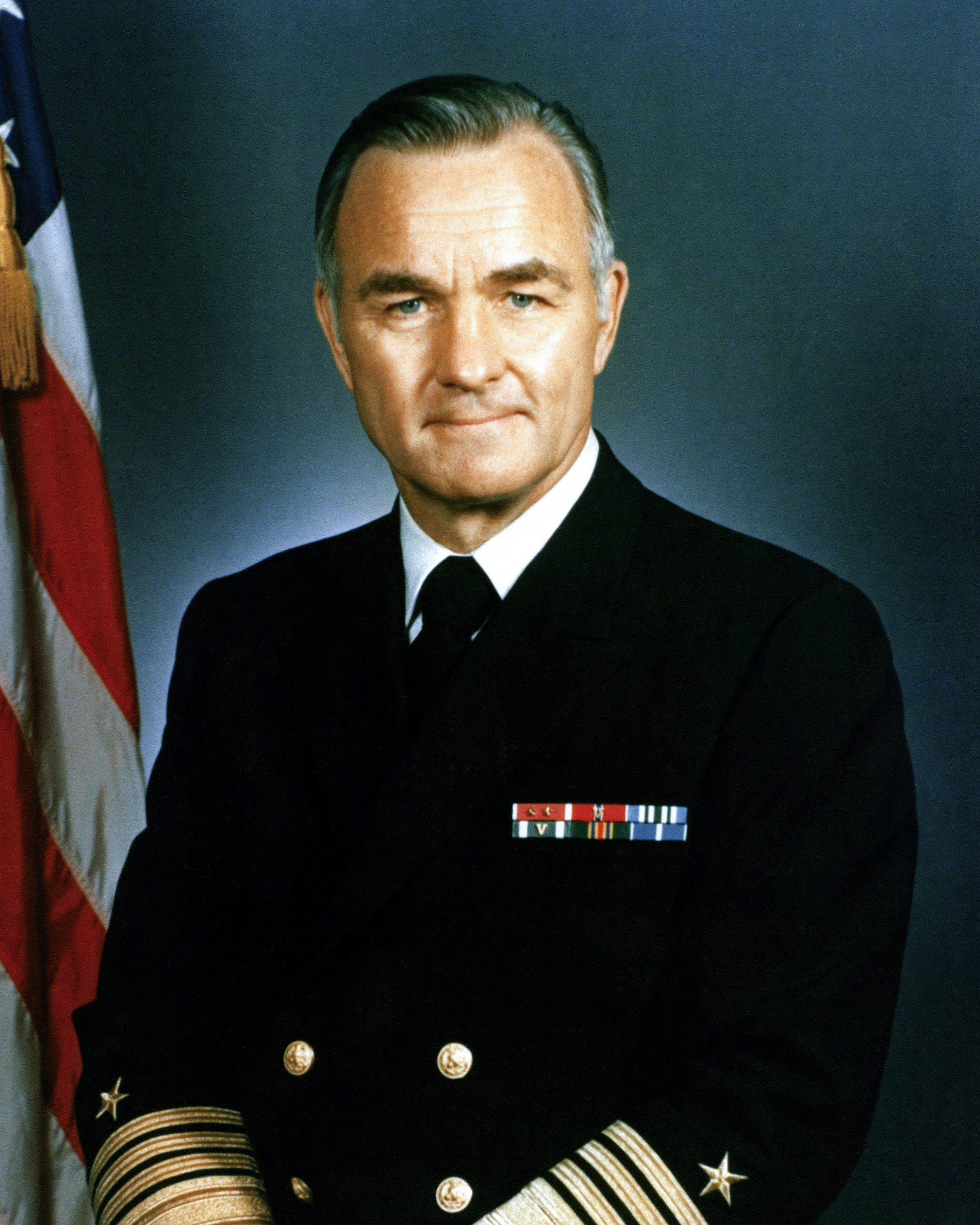 Admiral Stansfield Turner in a US Navy four-star dress uniform, ribbons on the breast and gold sleeve stripes at the cuff against the US flag, 1977