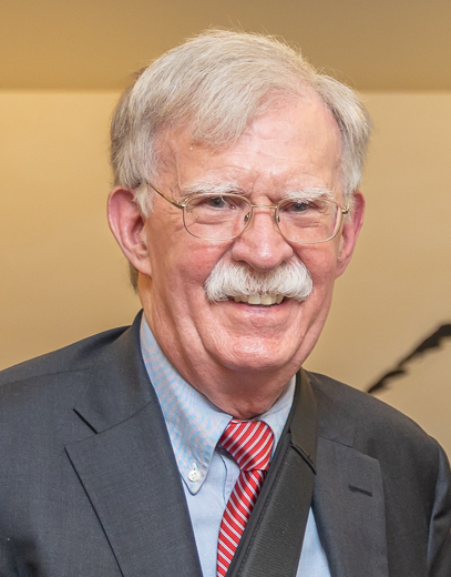 John Bolton smiling in a candid portrait, silver hair and thick mustache above a red-and-white striped tie under a grey suit, 2019