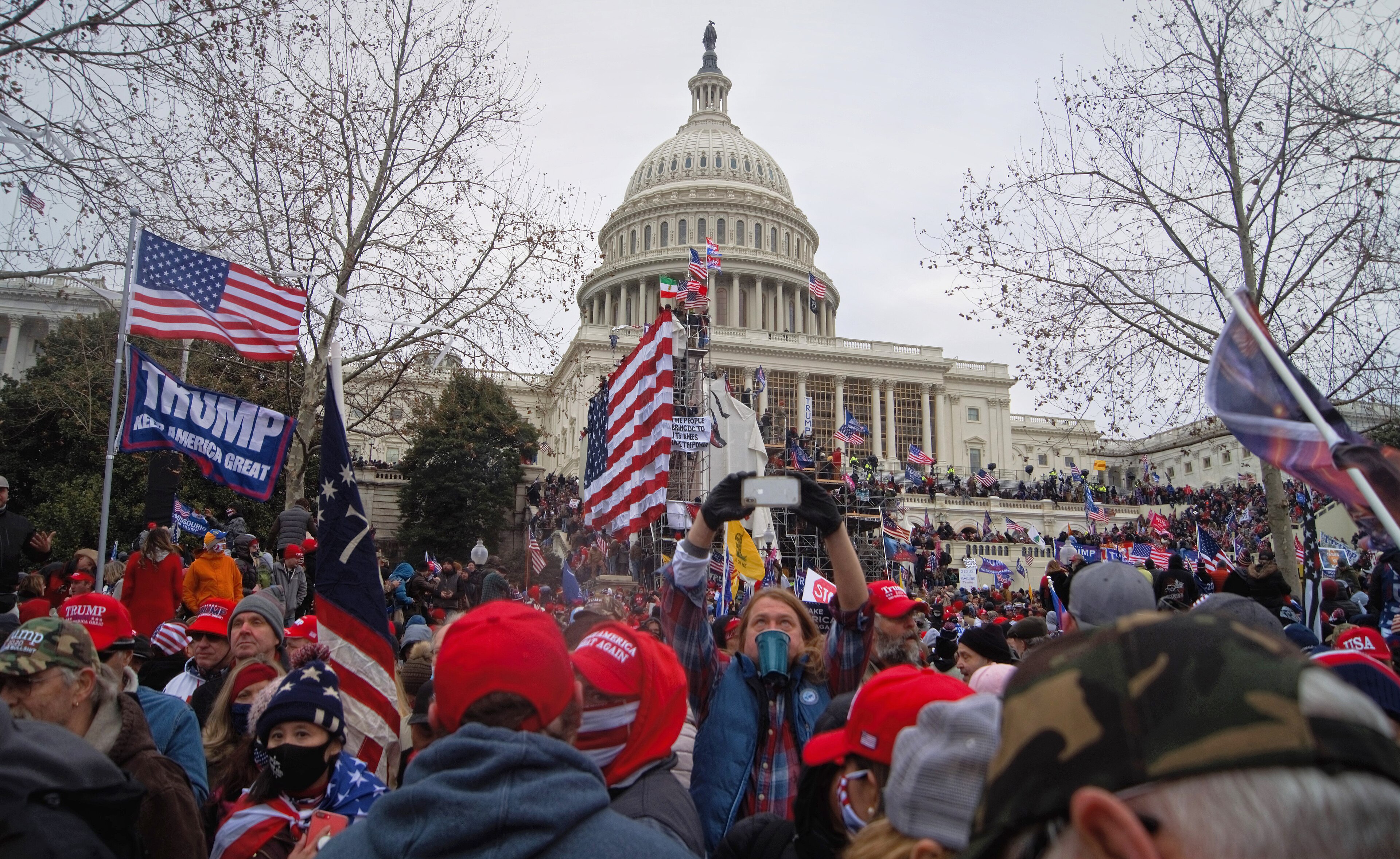 A dense crowd of Trump supporters in MAGA hats before the Capitol dome on January 6, flags and a Trump banner in the foreground, 2021