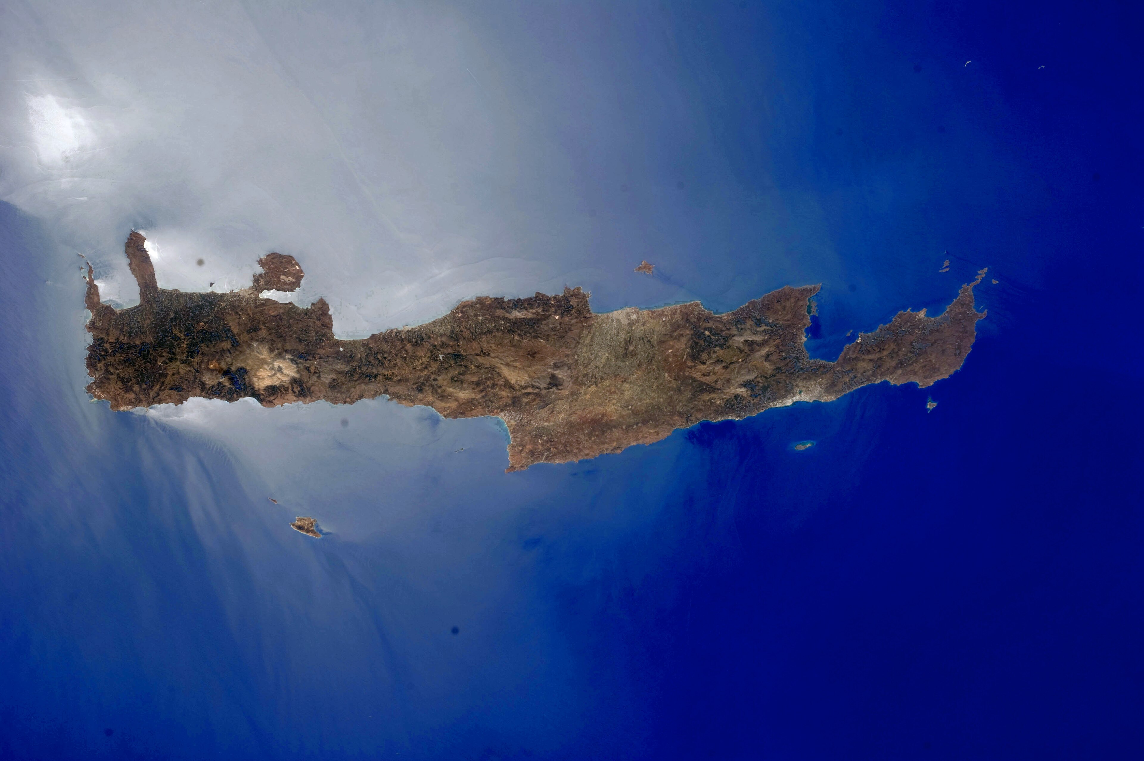 A satellite view of the elongated island of Crete against the blue Mediterranean, cloud banking off its northern coast, 2015