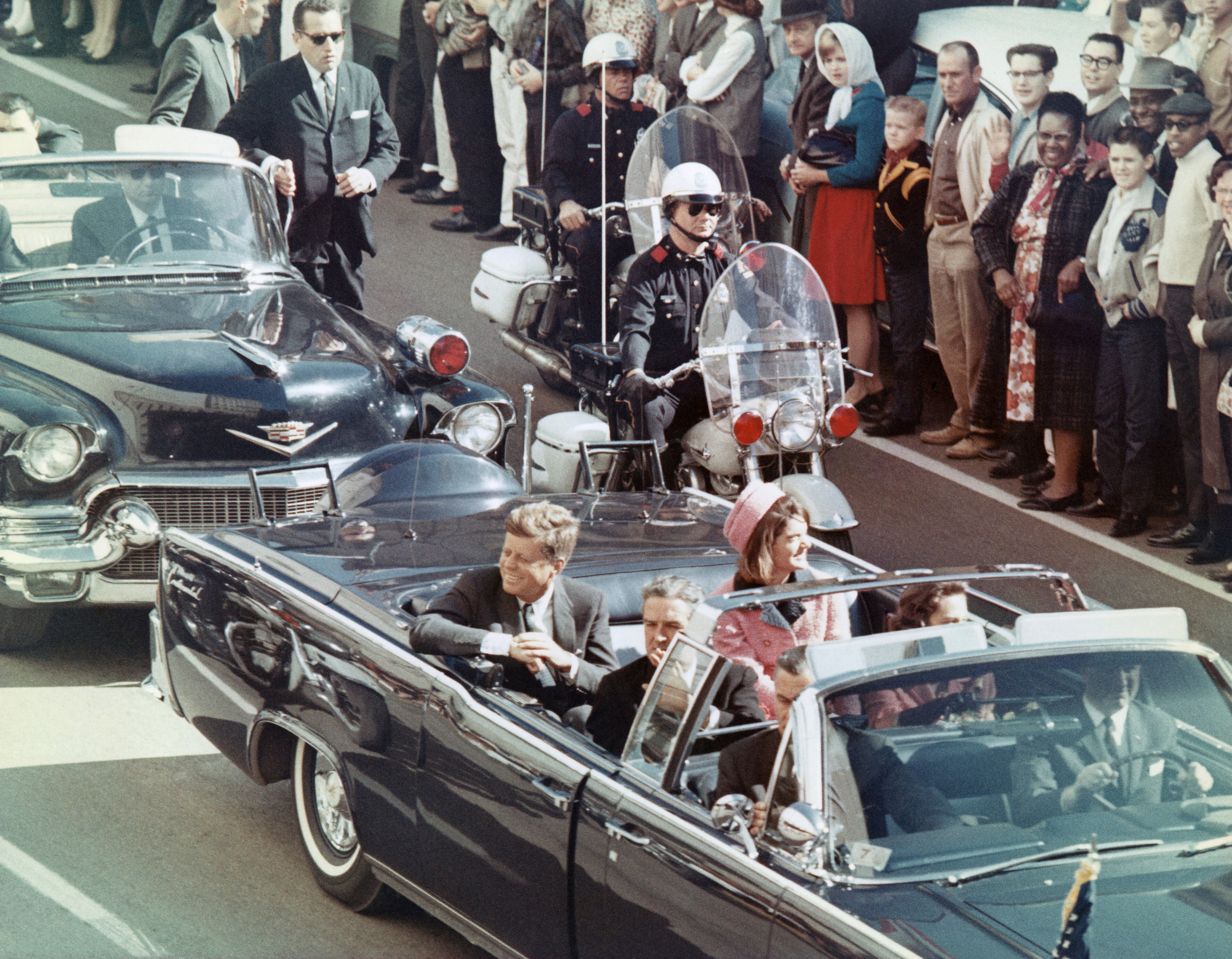John and Jackie Kennedy waving from an open limousine down Dallas' Elm Street beneath a motorcycle escort, crowds lining the curb, 1963