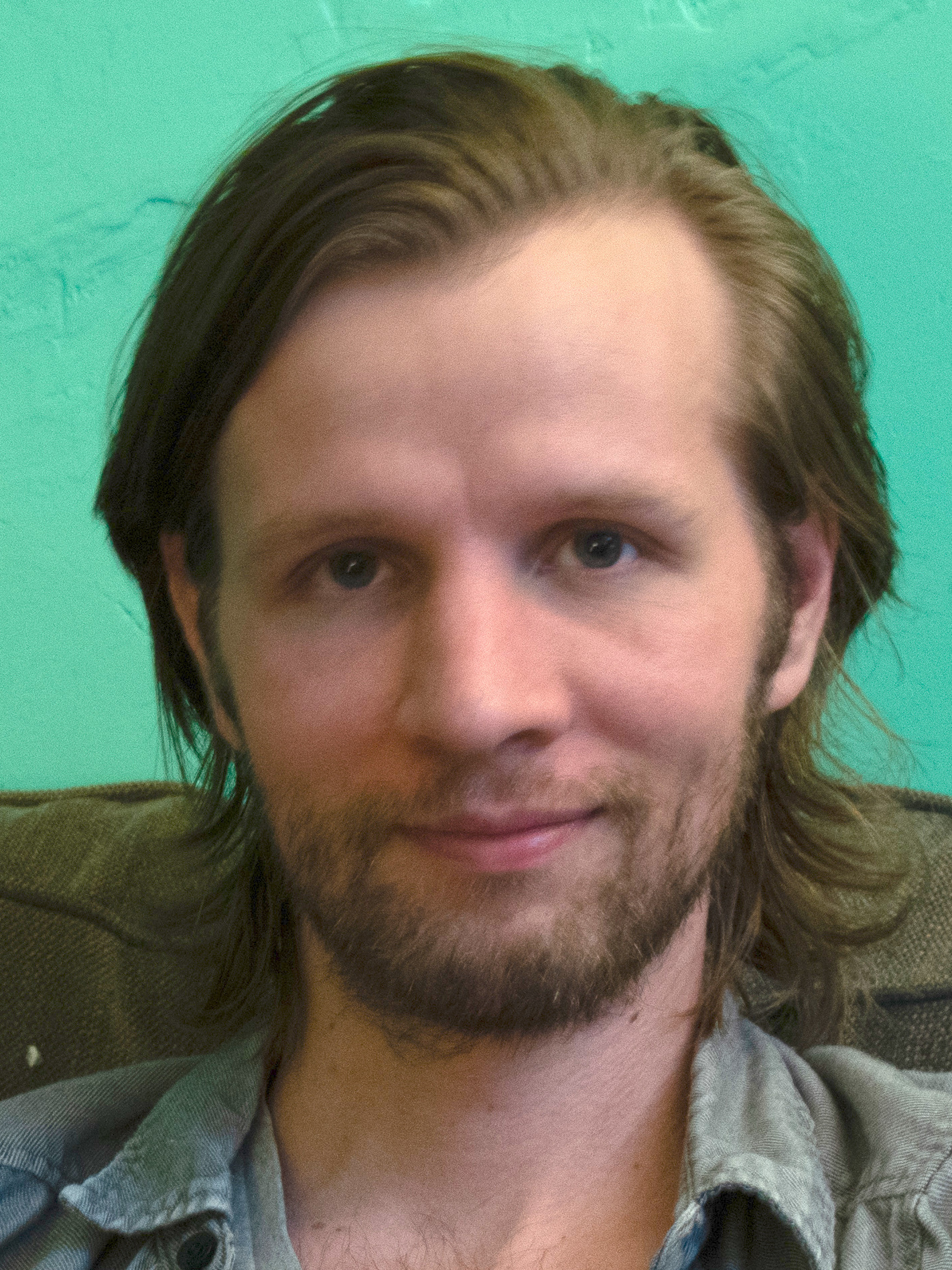 Daniel Hale in a candid portrait, long hair and beard over a grey shirt against a teal wall, 2019
