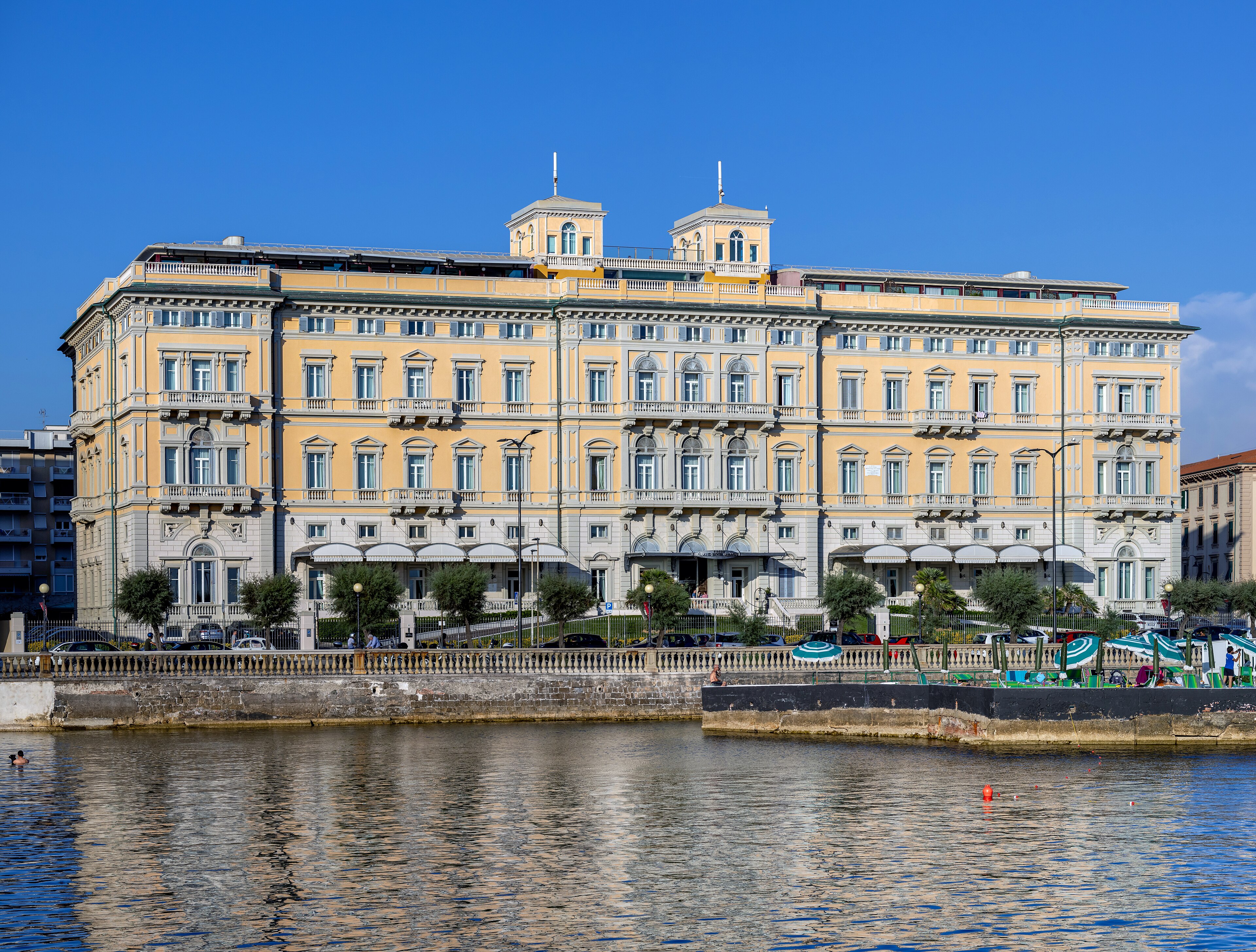 A yellow-and-white neoclassical waterfront hotel with twin cupolas at its roofline, seen across a calm Mediterranean harbour, 1990