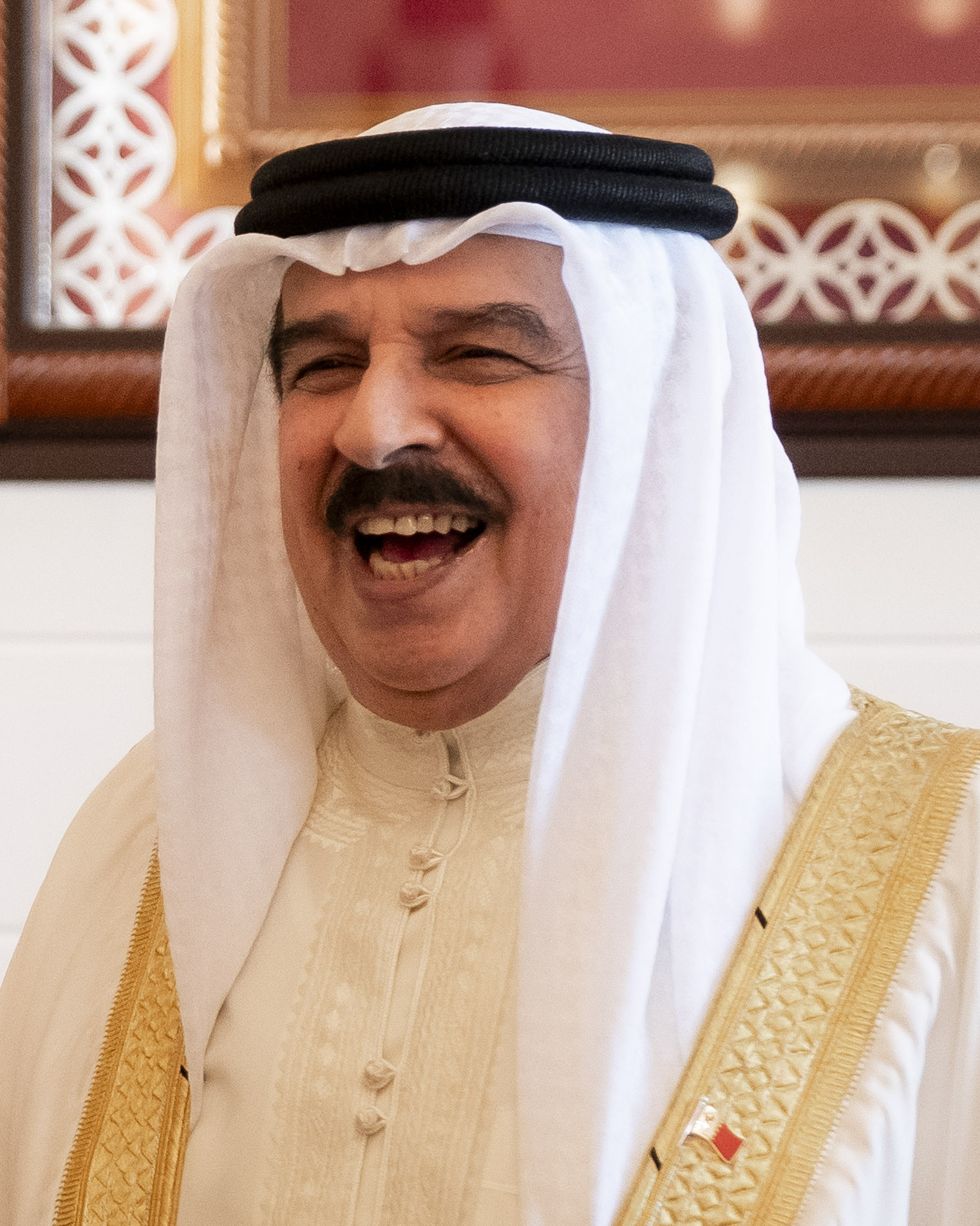 King Hamad bin Isa Al Khalifa laughing in a white ghutra and gold-trimmed bisht with a Bahraini flag pin on his lapel, 2018