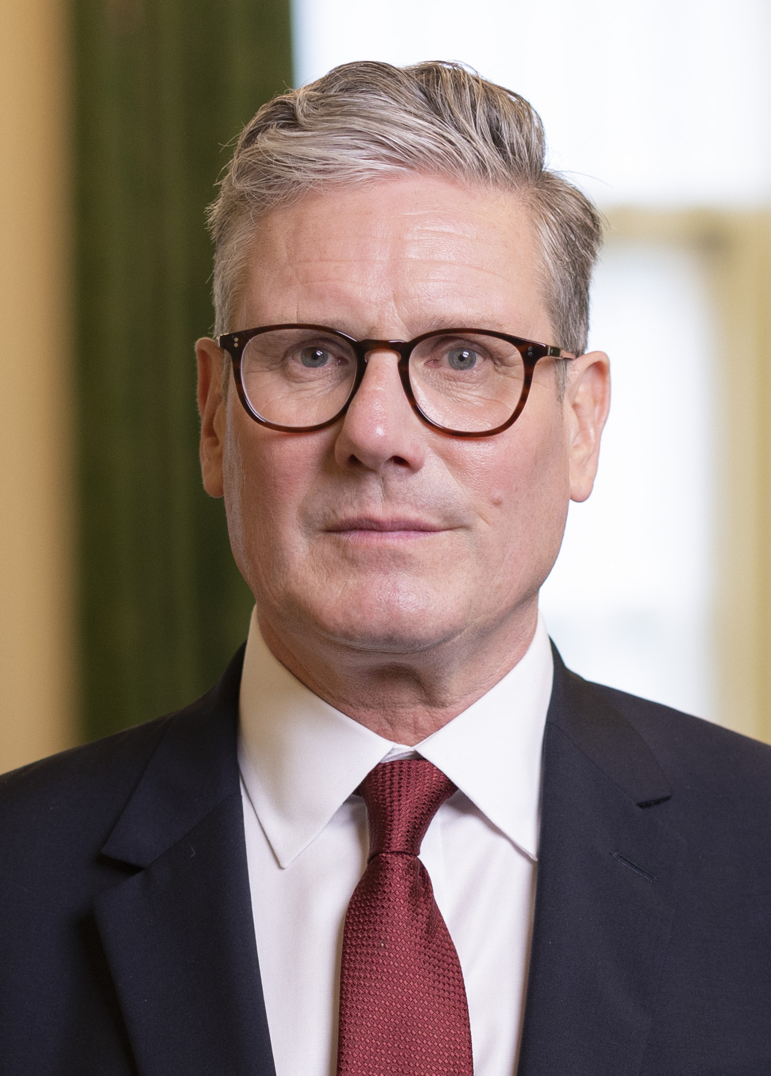 Keir Starmer in his official prime ministerial portrait, tortoise-shell glasses over a burgundy tie under a navy suit, 2024