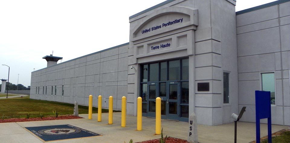 The entrance of the United States Penitentiary in Terre Haute, a low grey block flanked by yellow bollards and a distant guard tower, 2019
