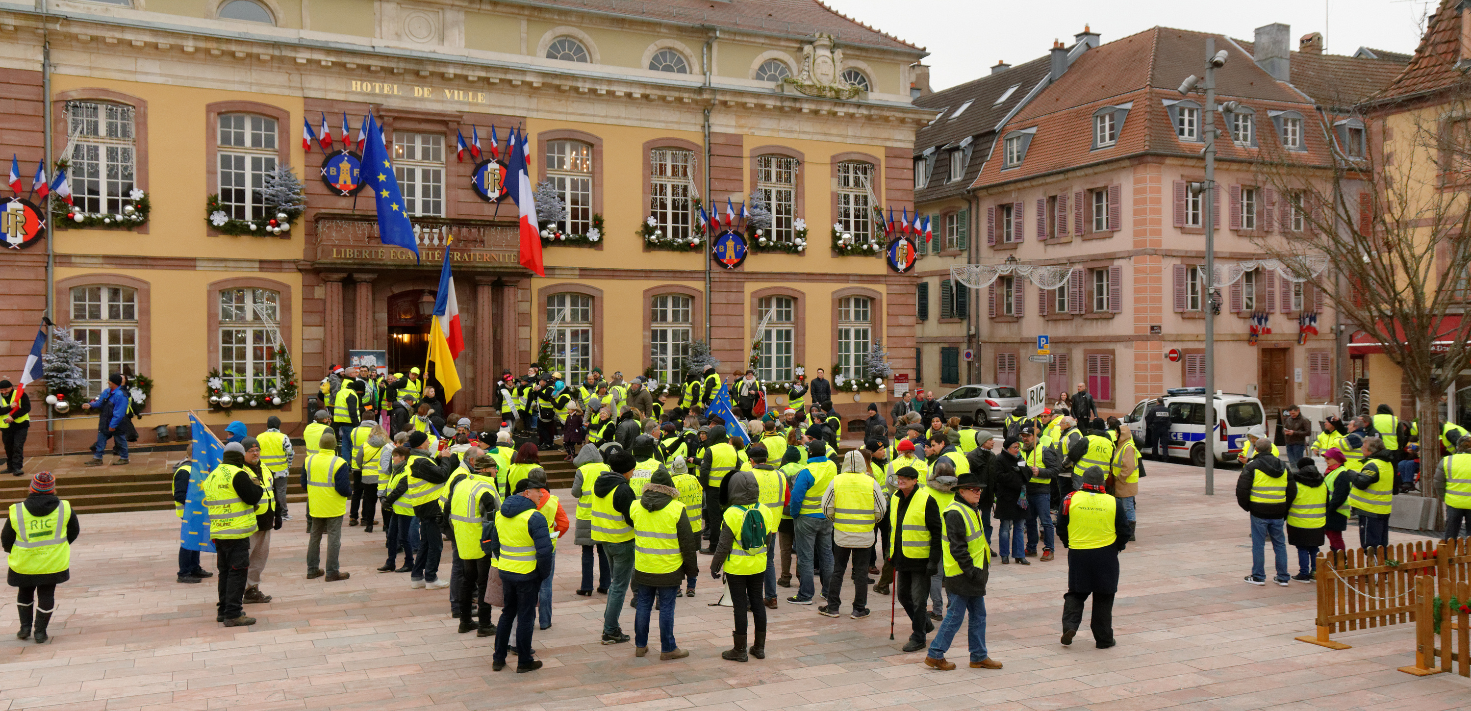 A gilets jaunes rally in the town square before a French Hôtel de Ville, protesters in high-visibility yellow vests packed under a facade hung with tricolor flags and Christmas garlands, 2018