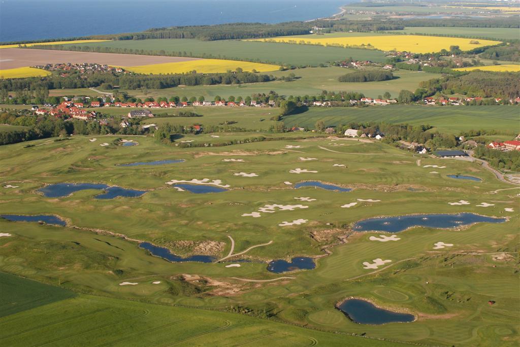 Aerial view of a Baltic coast golf course, kidney-shaped ponds and white sand bunkers set among green fairways above a rapeseed-flowered plain, 2015