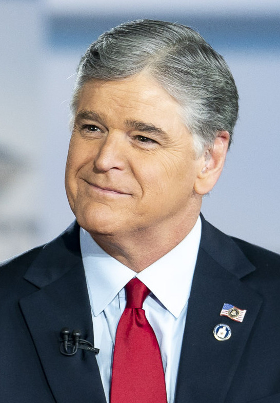 Sean Hannity smiling on a Fox News set in a navy blazer over a white shirt and red tie, US-and-Trump lapel pins on his lapel, 2018